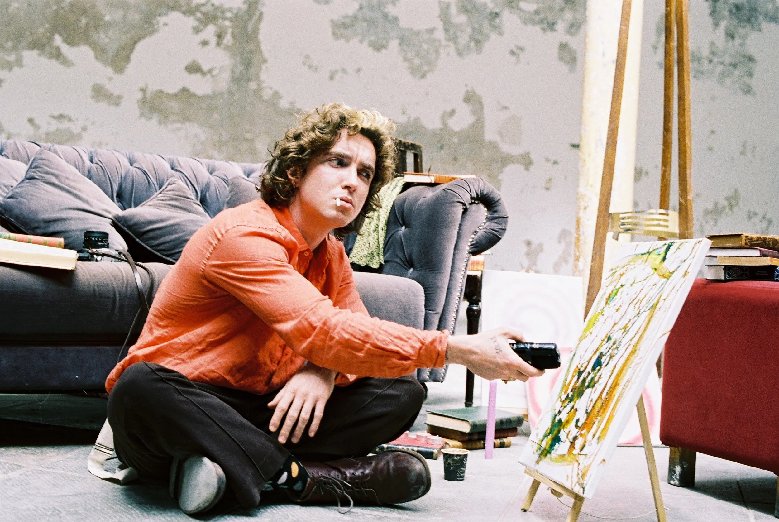 A woman with curly hair wearing an orange shirt is sitting on the floor, holding a remote control, and looking at a canvas with abstract paint. There are books and art supplies around her, and a gray couch behind her.