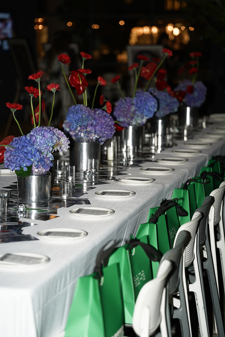 Elegant dining table set for a formal event with floral centerpieces featuring blue hydrangeas and red anthuriums in metallic vases, white plates, silverware, small glass candle holders, green gift bags hanging on chairs, and white chairs, in a warml