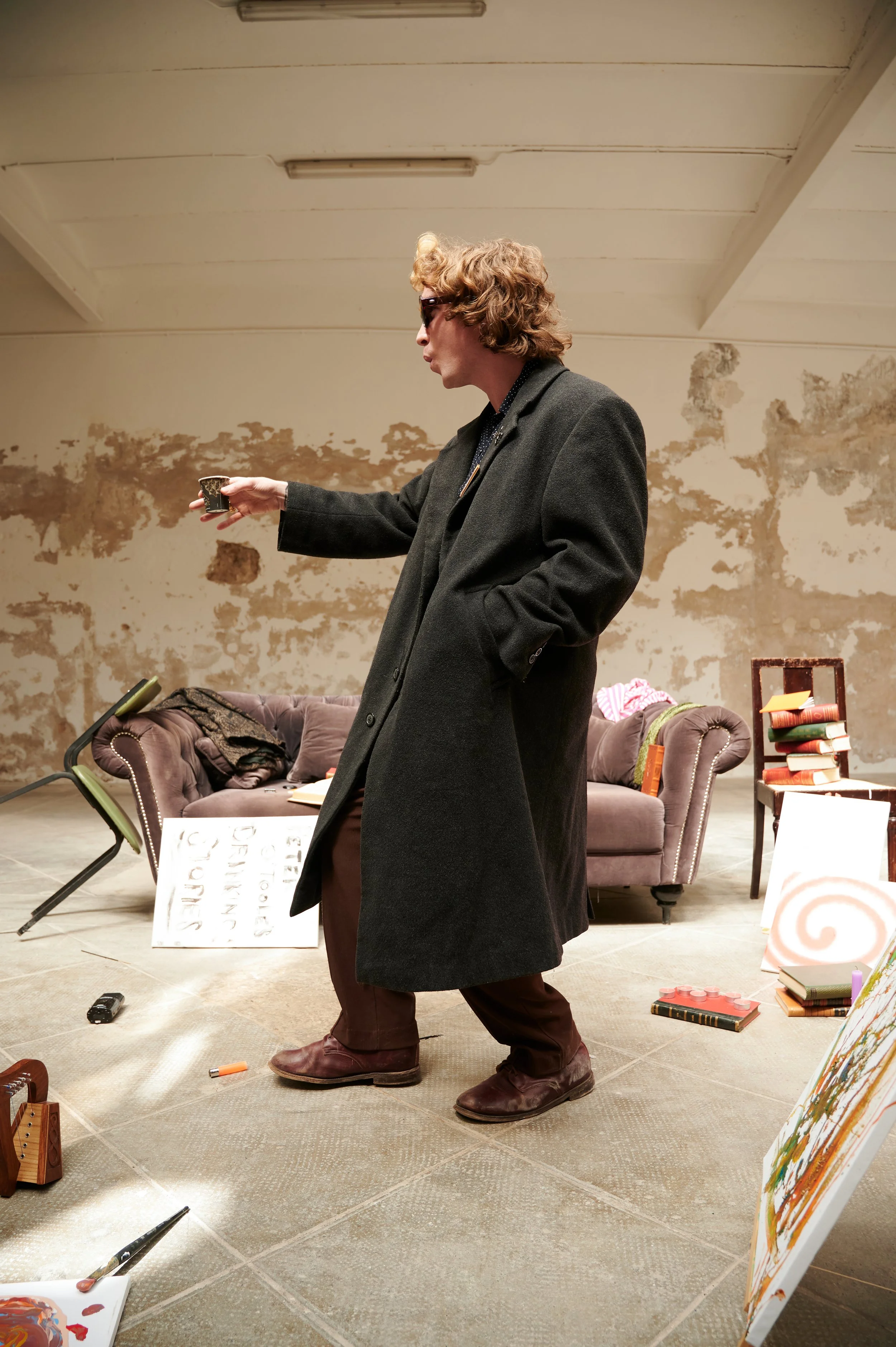A person with curly blonde hair wearing sunglasses, a black coat, and brown pants is holding a small cup and walking in a room with a distressed wall background, scattered art supplies, a sofa with clothing and bags, and art canvases.