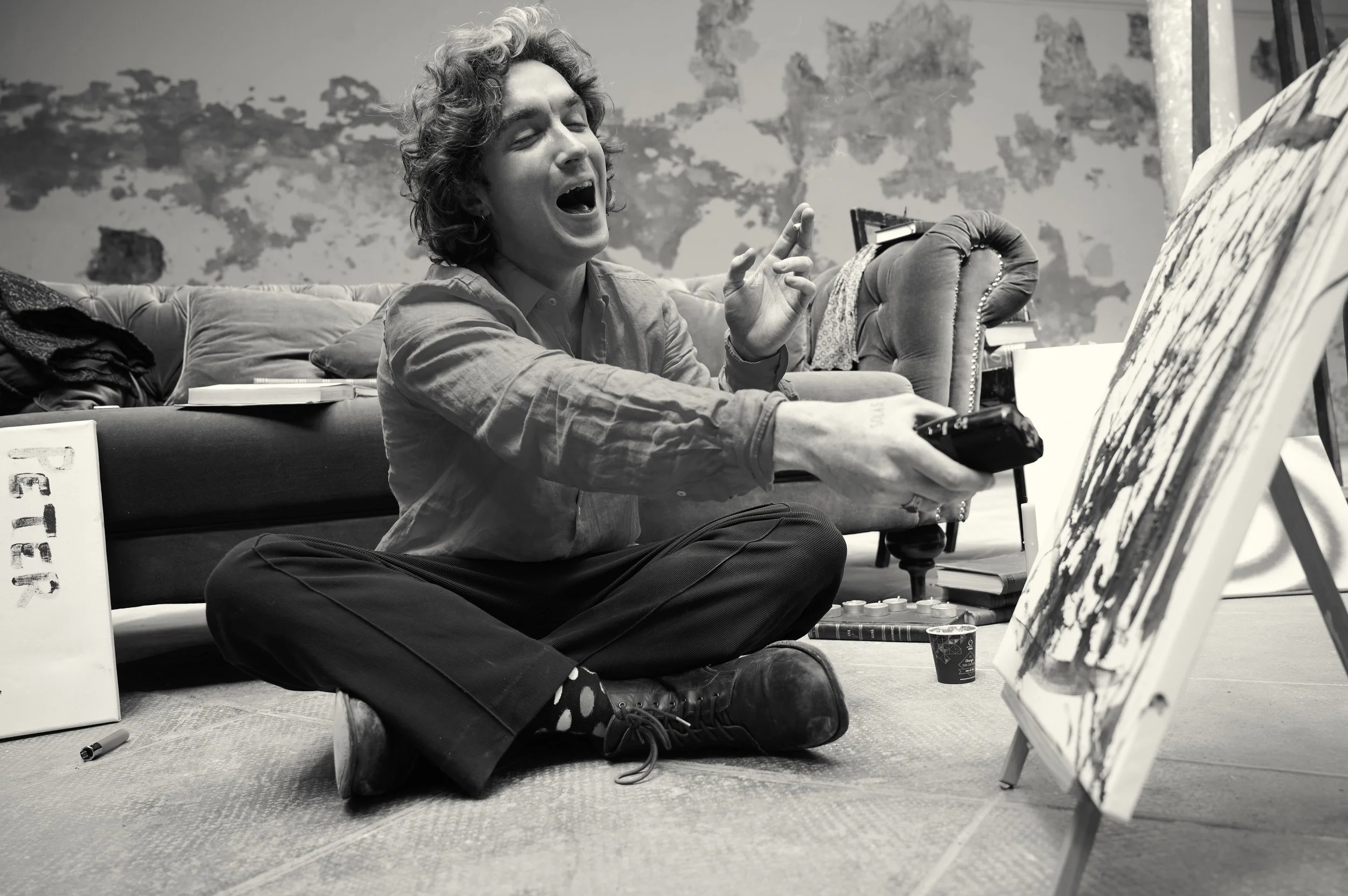 A person with curly hair sitting cross-legged on the floor, singing or speaking passionately, while holding a remote control. They are surrounded by art supplies, including a canvas with abstract painting, books, and a cup, in a room with a textured 