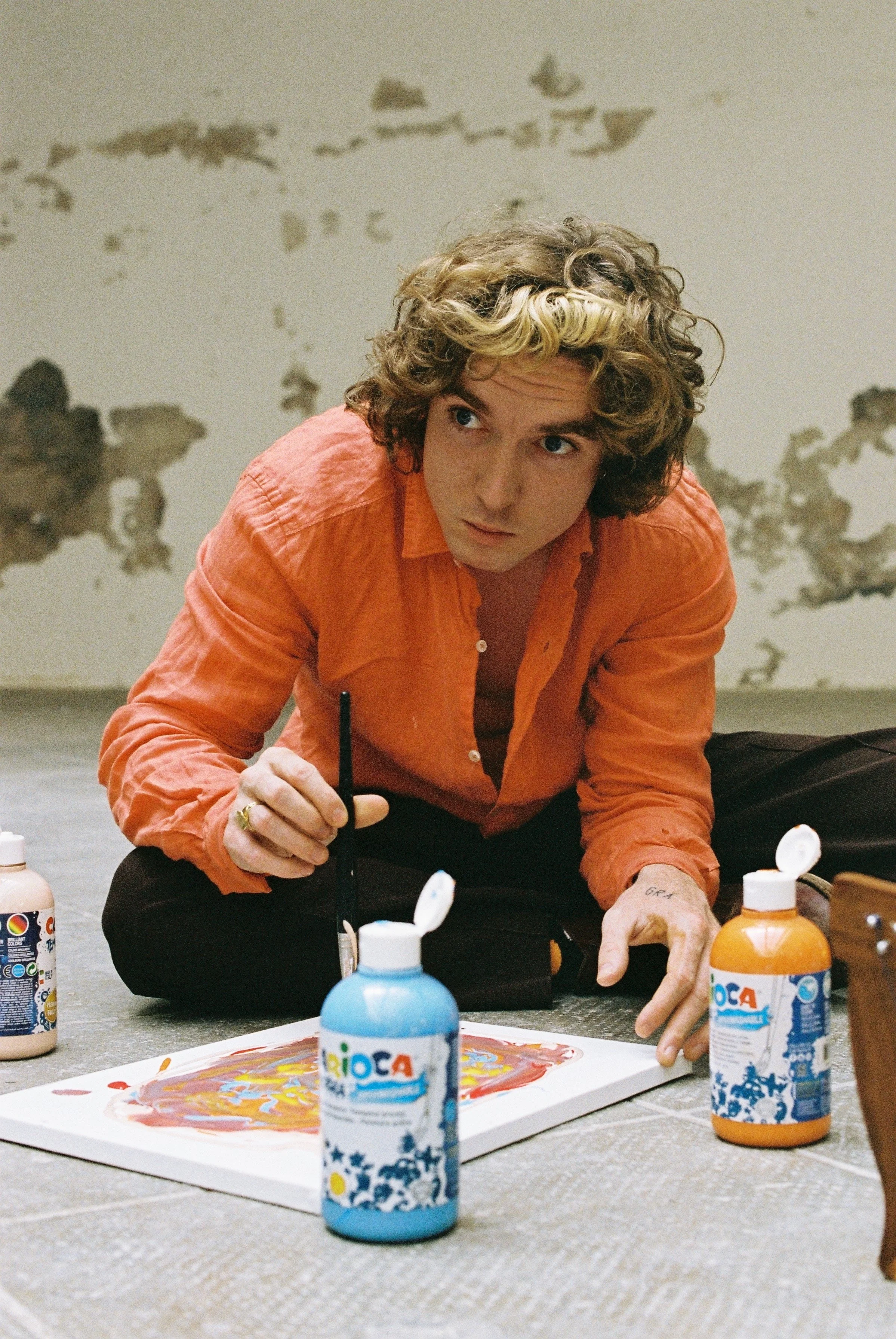 A young man with curly blonde hair wearing an orange shirt, sitting cross-legged on the floor, painting a colorful design on a canvas. Several bottles of colorful paint are nearby.