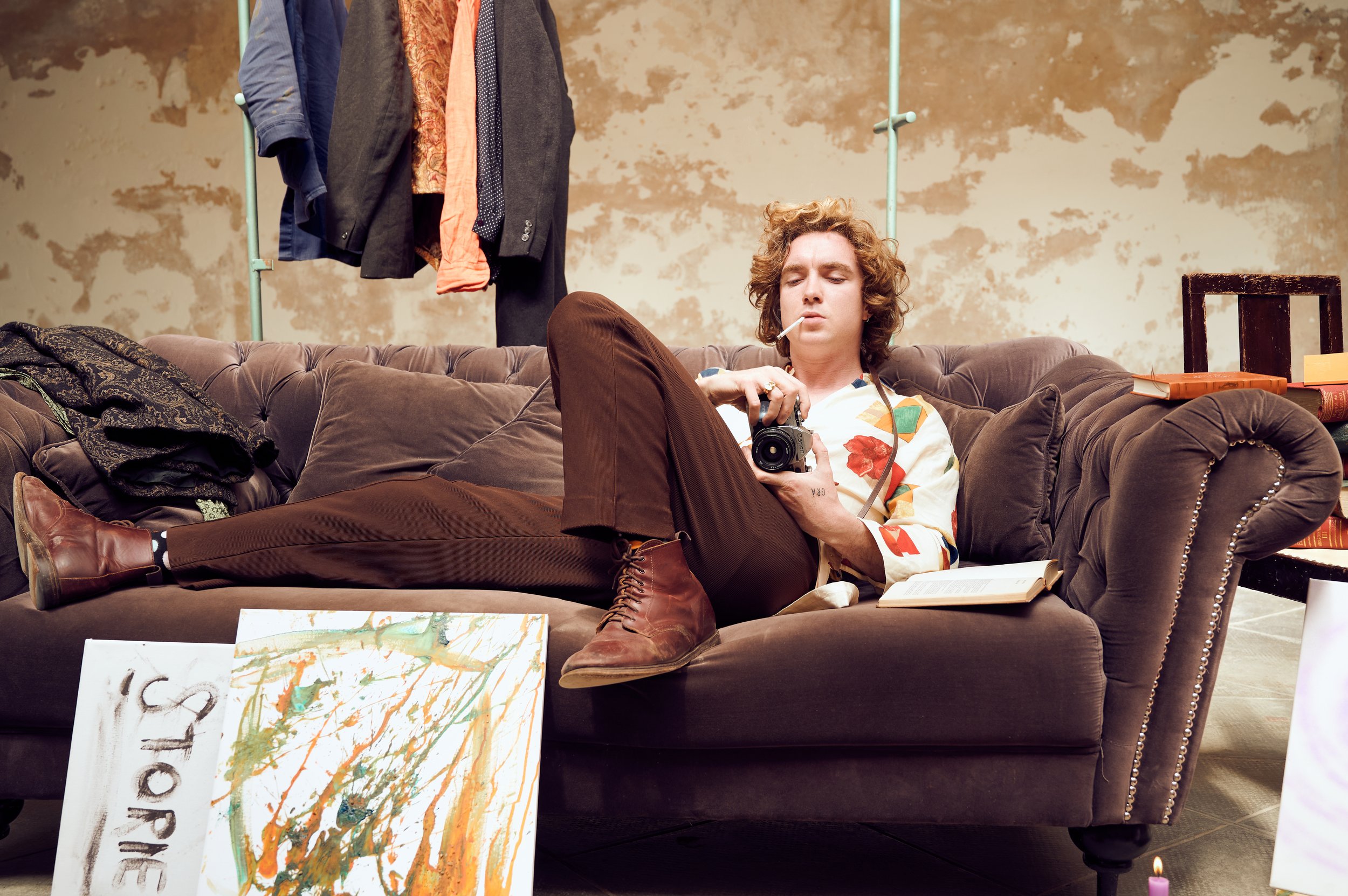 A young man with curly hair reclines on a vintage brown velvet couch, smoking a cigarette, holding a camera, and reading a book. Behind him is a rustic wall with peeling paint, and a clothing rack with various jackets and shirts. In front of the couc