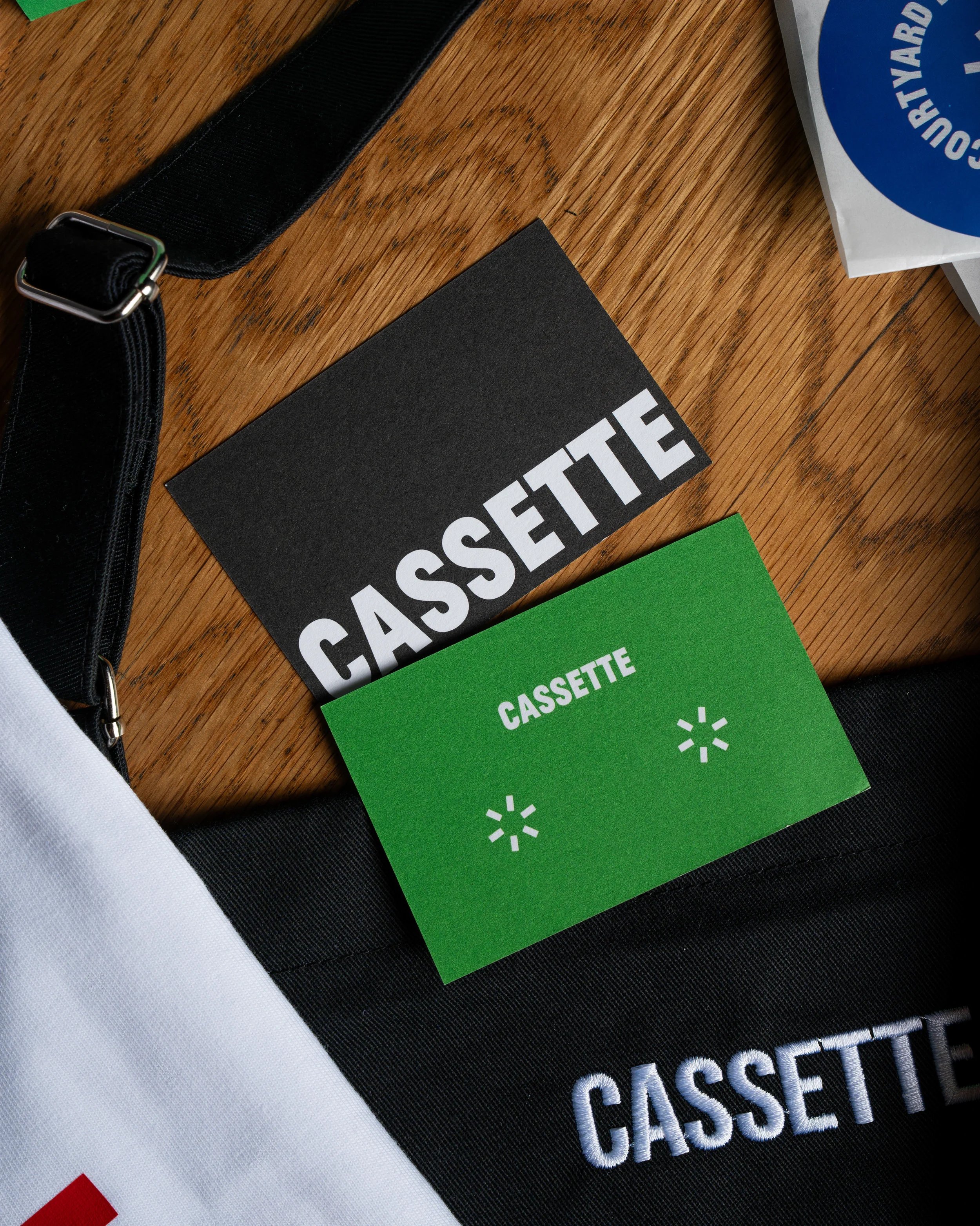 Close-up of a black bag with white embroidered logo, placed on a wooden surface with cassette tapes and colorful cards.