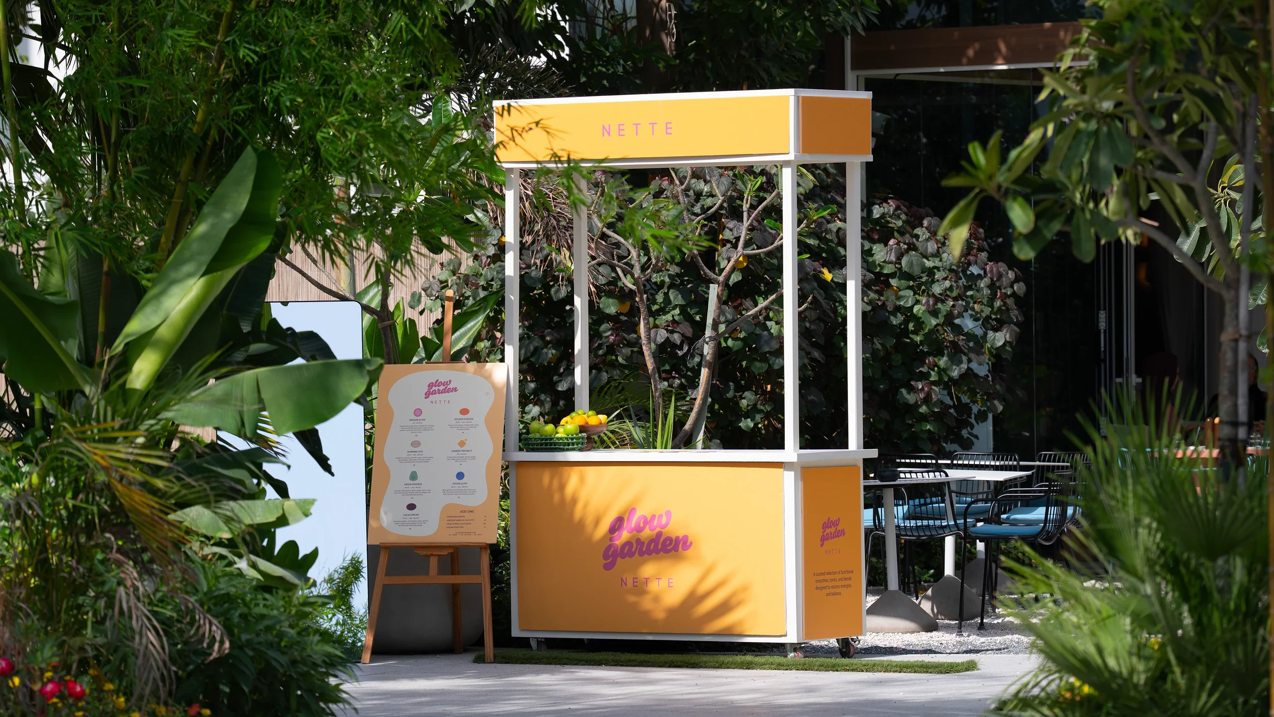 A yellow and white kiosk with the words "Glow Garden" and "NETTE" on it, surrounded by lush green plants, with a menu sign and a bowl of green and orange fruits.