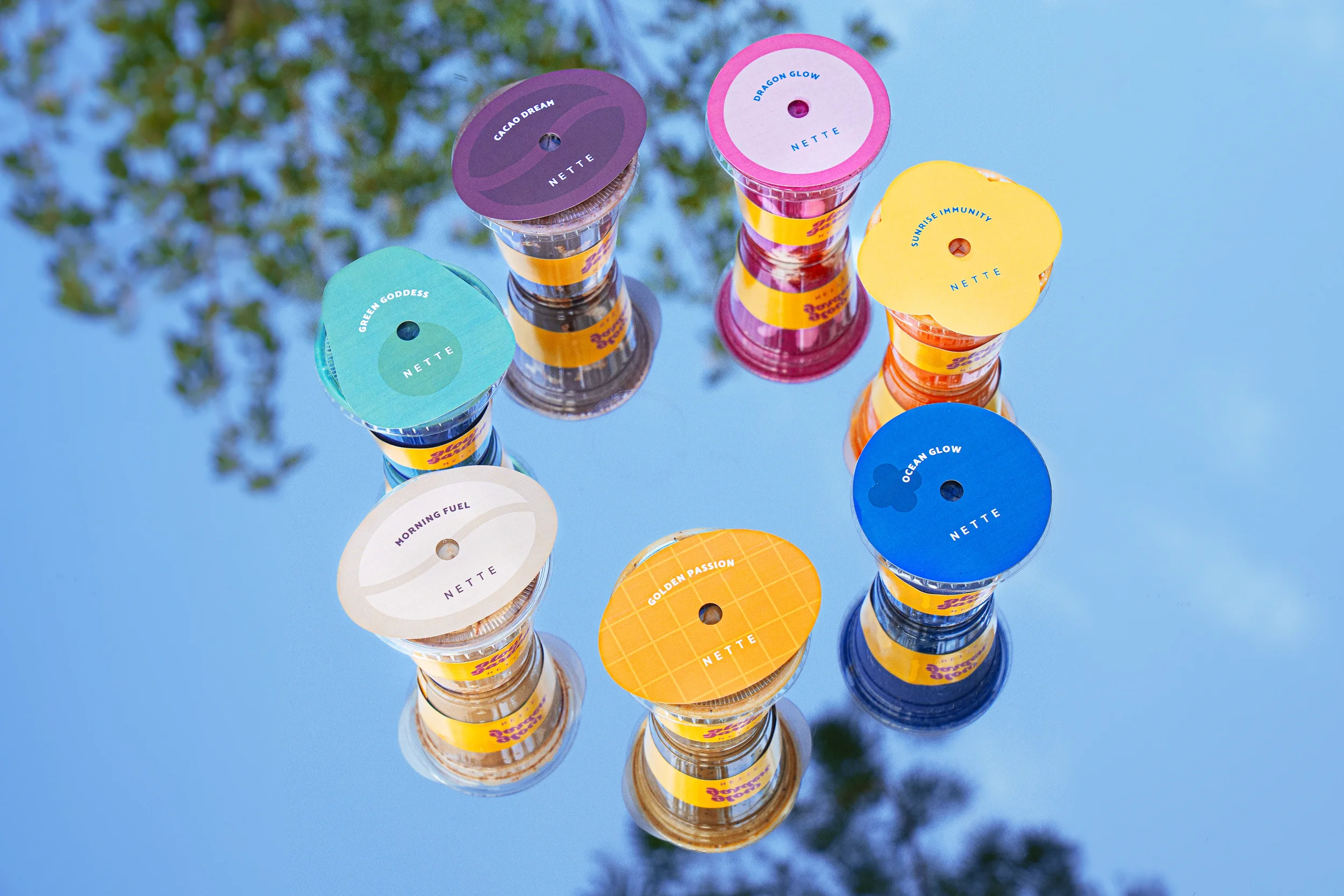 Multiple stacks of small bottles with colorful labels on a reflective surface, with a blurred green tree and blue sky in the background