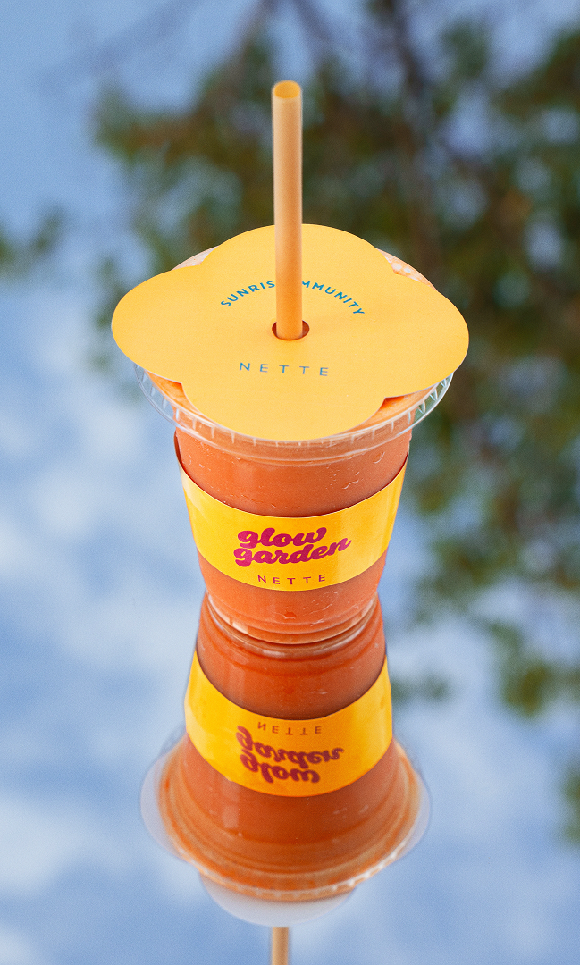 A cold beverage in a clear plastic cup with a yellow paper cover and a straw. The cup has a yellow label with pink text that reads 'Glow Garden Nette.' The cup is reflected in a shiny surface, with a blurred background of trees and sky.