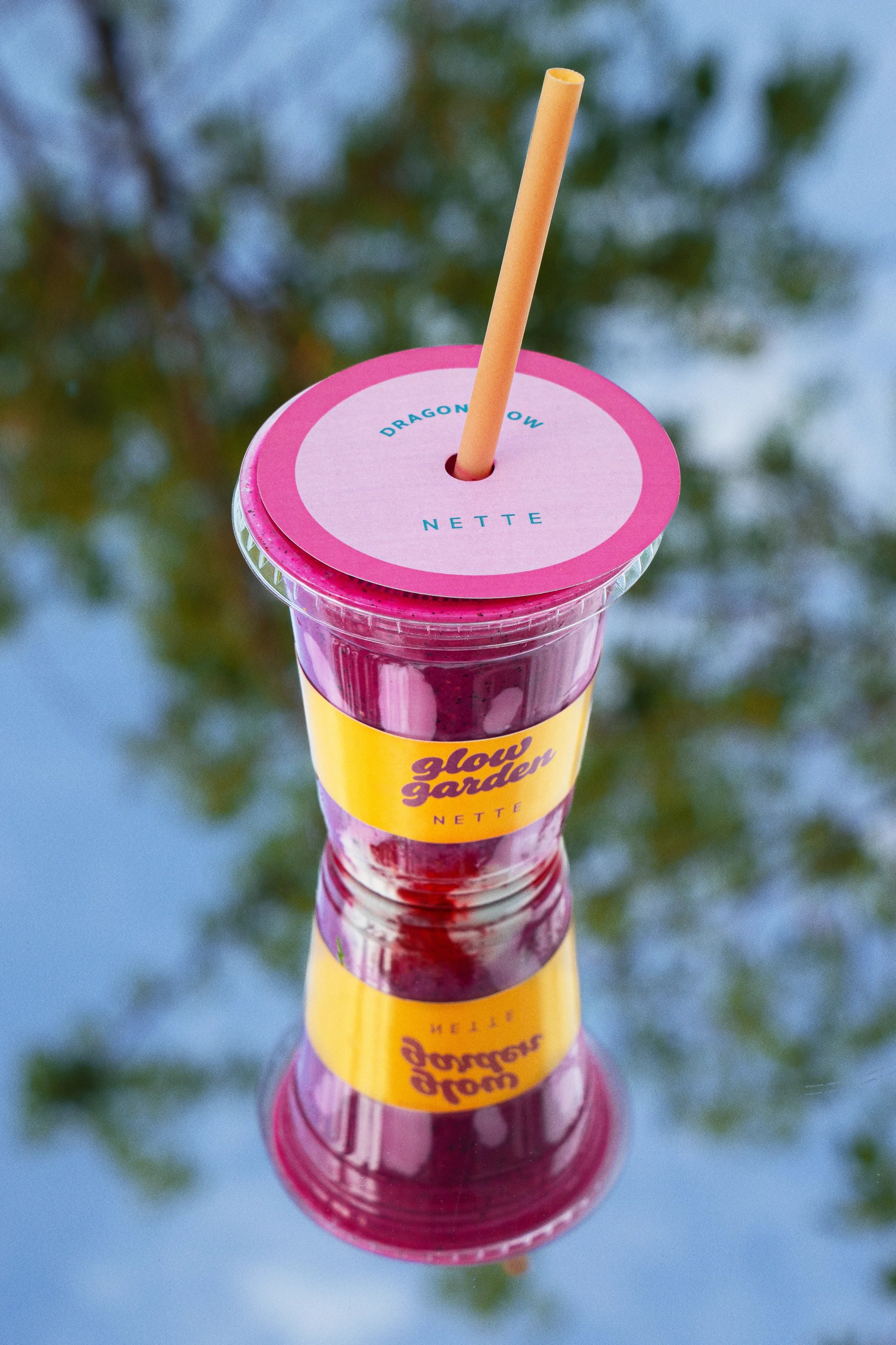 A plastic bubble tea drink with a pink straw and a pink and yellow label that reads "Glow Garden" and "NETTE." The drink appears to be purple in color and is placed on a reflective surface that mirrors the drink. The background is blurred with green 