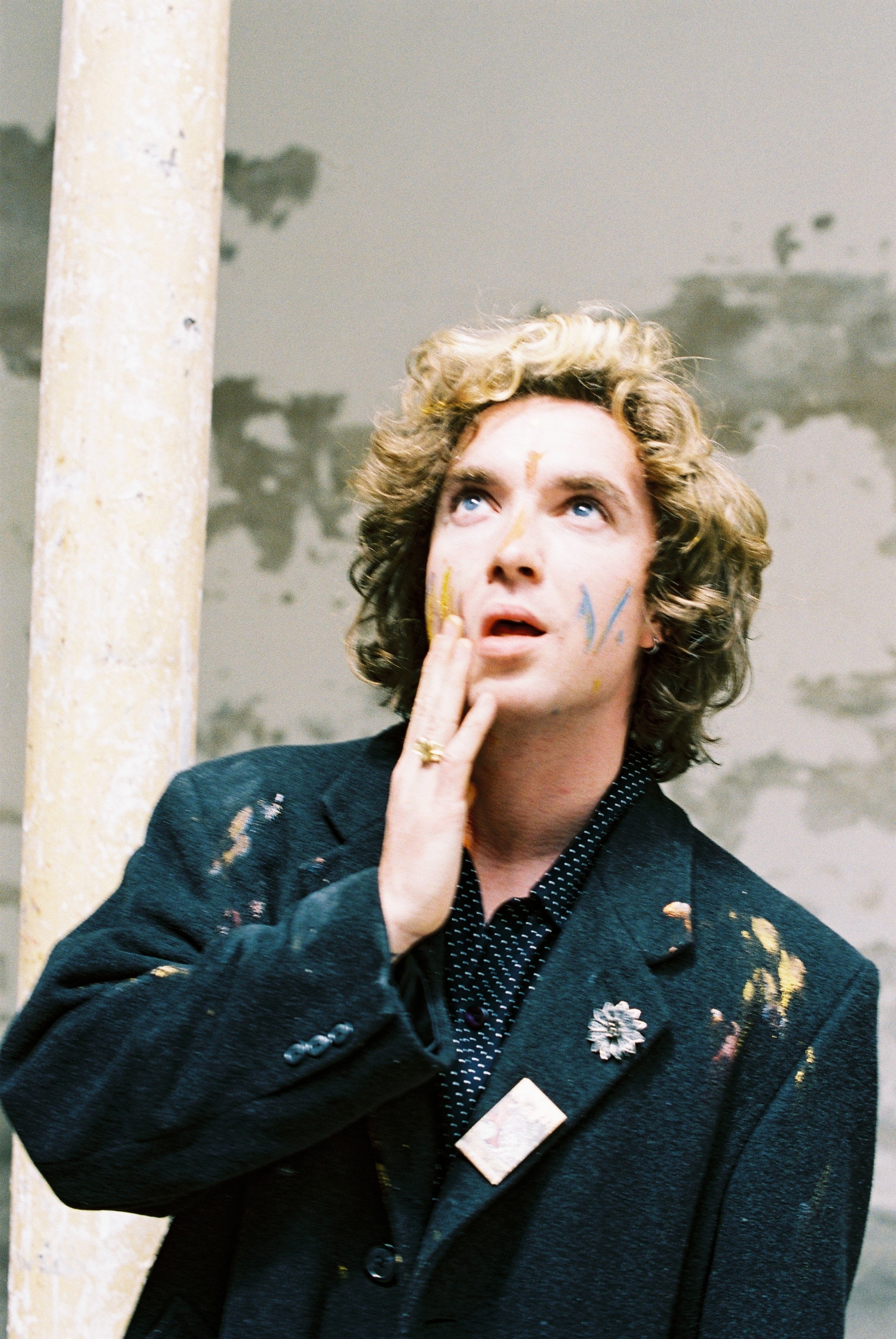 A person with curly blonde hair and blue eyes, wearing a dark blazer with paint splatters, a polka dot shirt, and a flower-shaped pin, touching their lips with their fingers, standing in front of a wall with a pipe and a tree shadow.