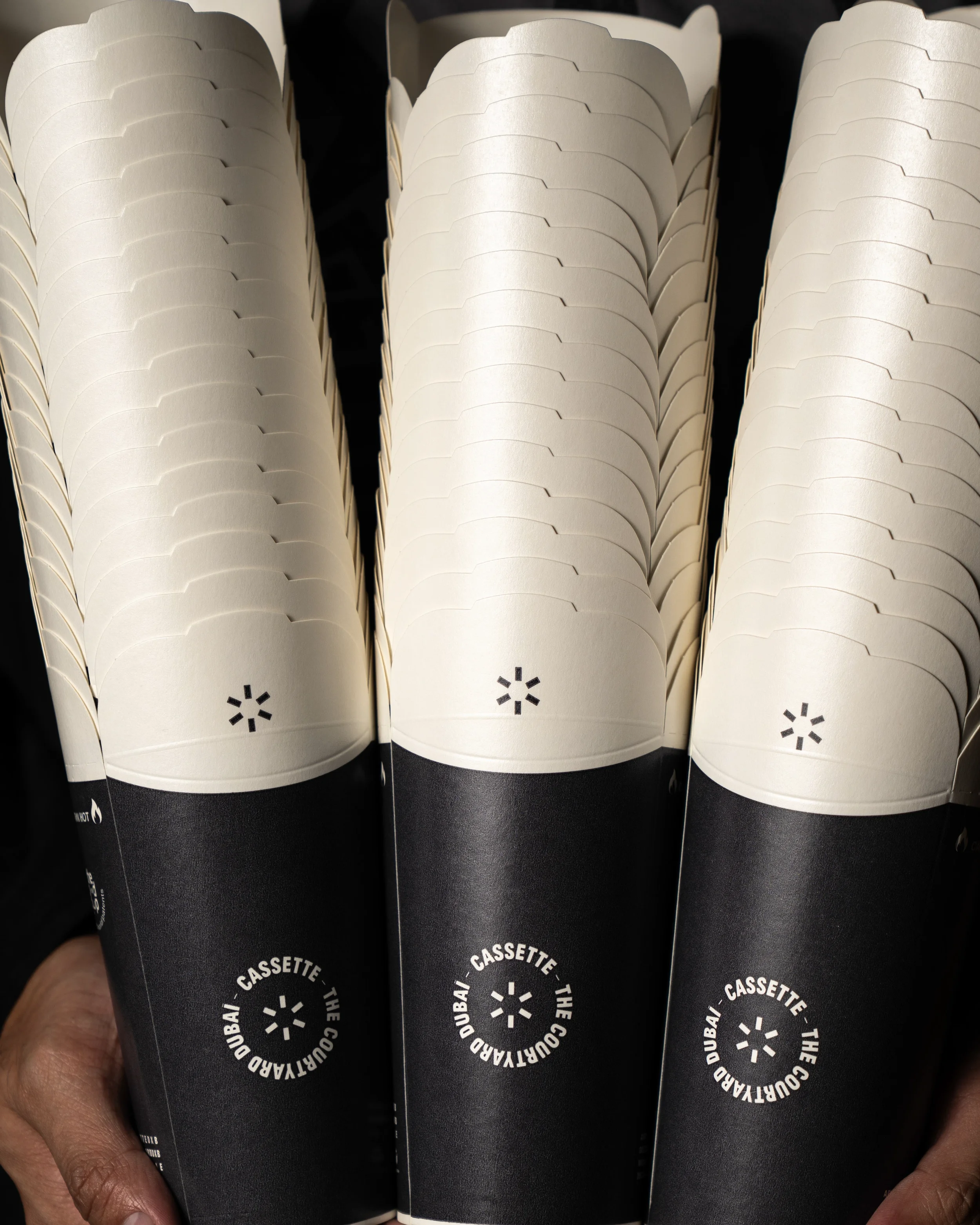 A hand holding several black and white paper cups with a circular logo and text that reads "Dubai - Cassette The".