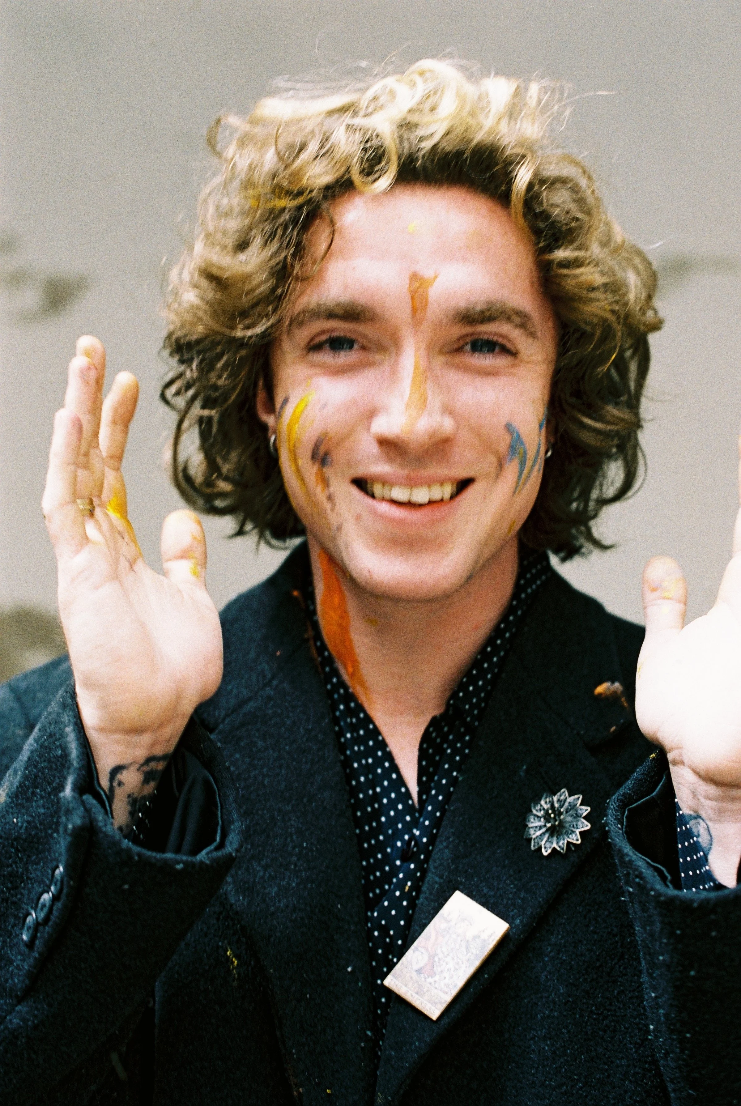 A person with curly blonde hair, painted face, and hands raised, smiling at the camera, wearing a dark jacket and polka dot shirt.