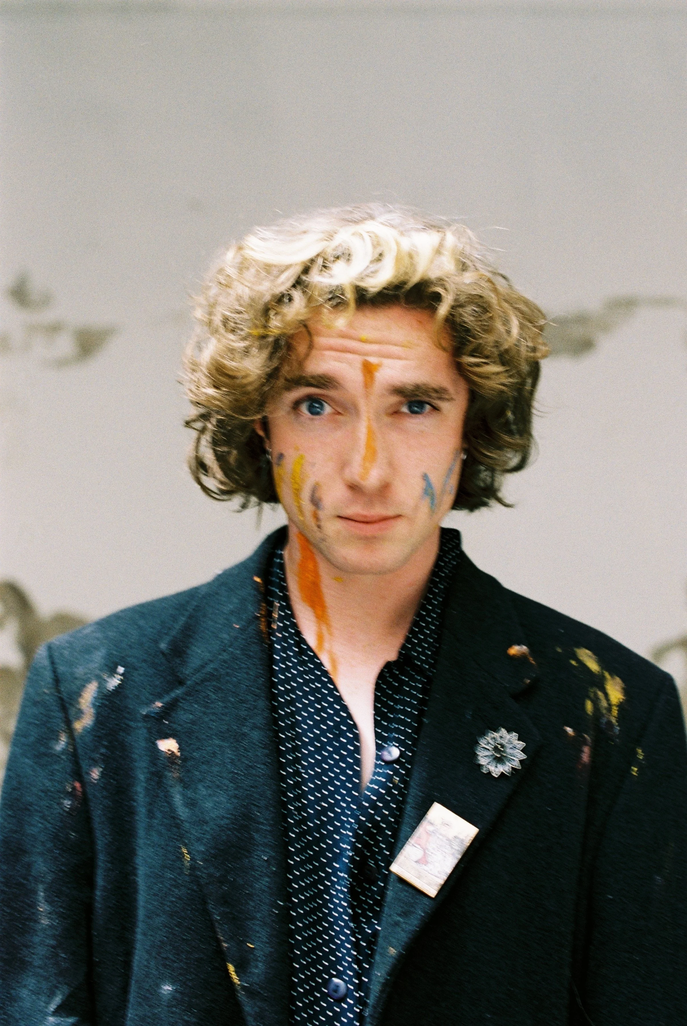 A young man with curly blonde hair and blue eyes, wearing a dark textured jacket with a brooch on the lapel, a polka dot shirt, and a name badge, stands in front of a white background with ceiling fans.