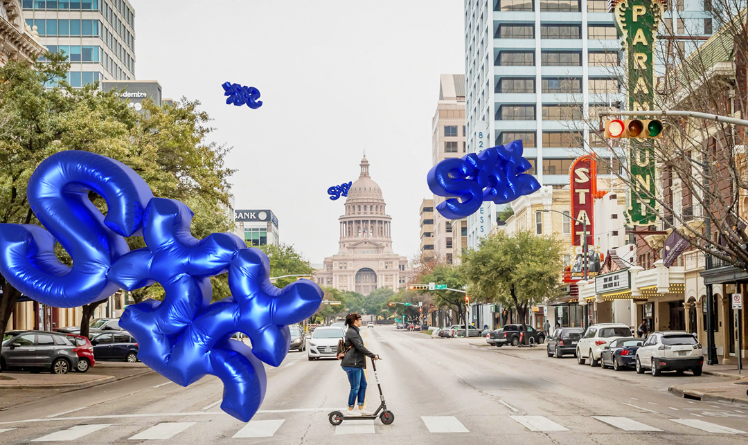 Street scene in downtown Texas with people, cars, and large blue balloon sculptures spelling 'TX', 'SALE', and other words, with the Texas State Capitol building in the background.