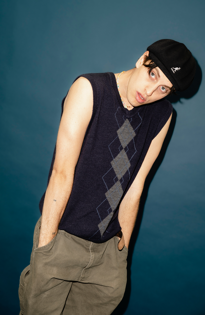 A young man with pale skin, black hair, and light-colored eyes wearing a black beanie hat, a sleeveless dark sweater with a diamond pattern, beige cargo pants, and a beaded necklace. He poses against a plain blue background, leaning slightly with han