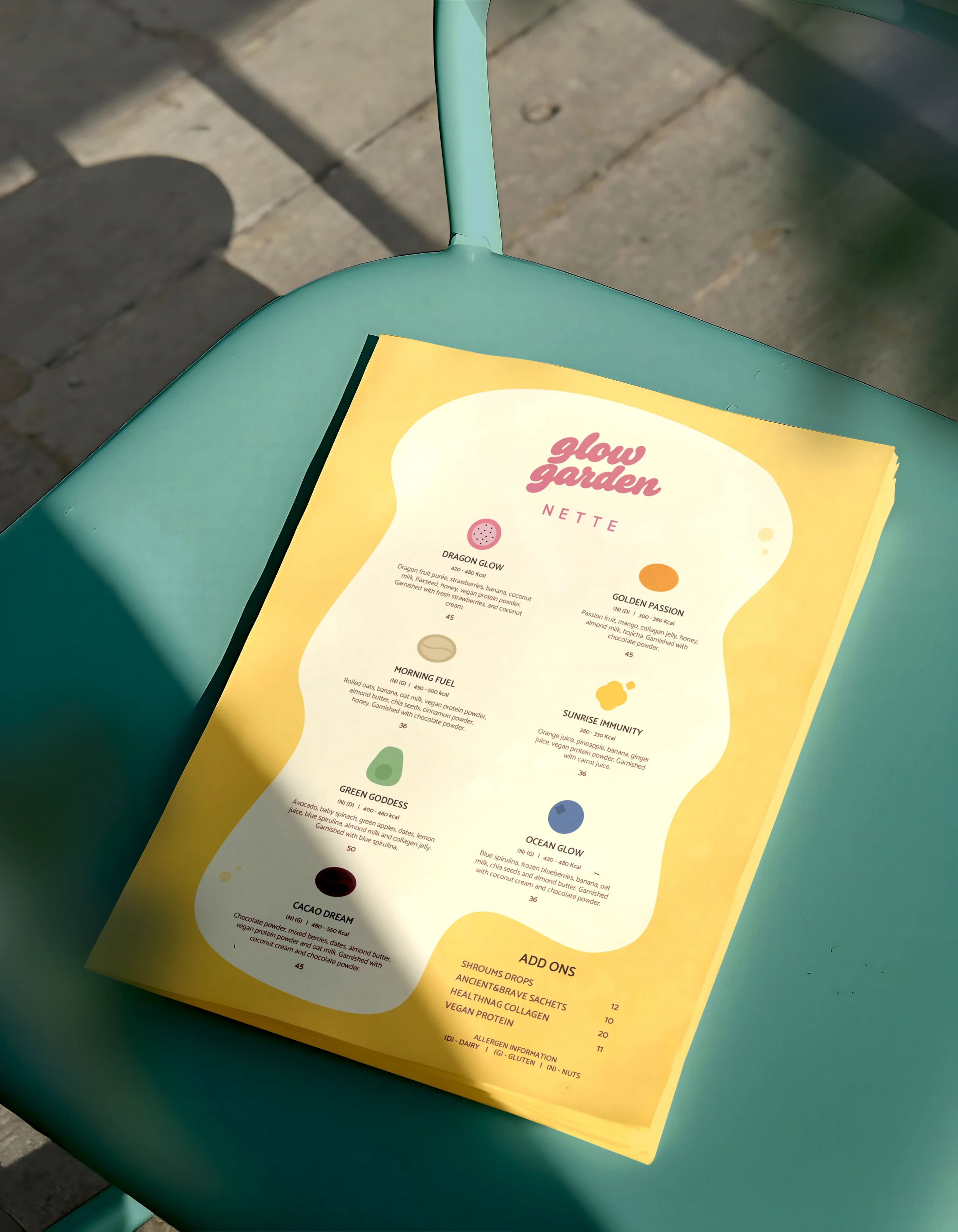 A menu featuring various smoothie flavors such as Glow Garden, Dragon Glow, Golden Passion, Morning Fuel, Sunrise Immunity, Green Goddess, Ocean Glow, Cacao Dream, and Add Ons, placed on a teal outdoor chair with a concrete pavement background.