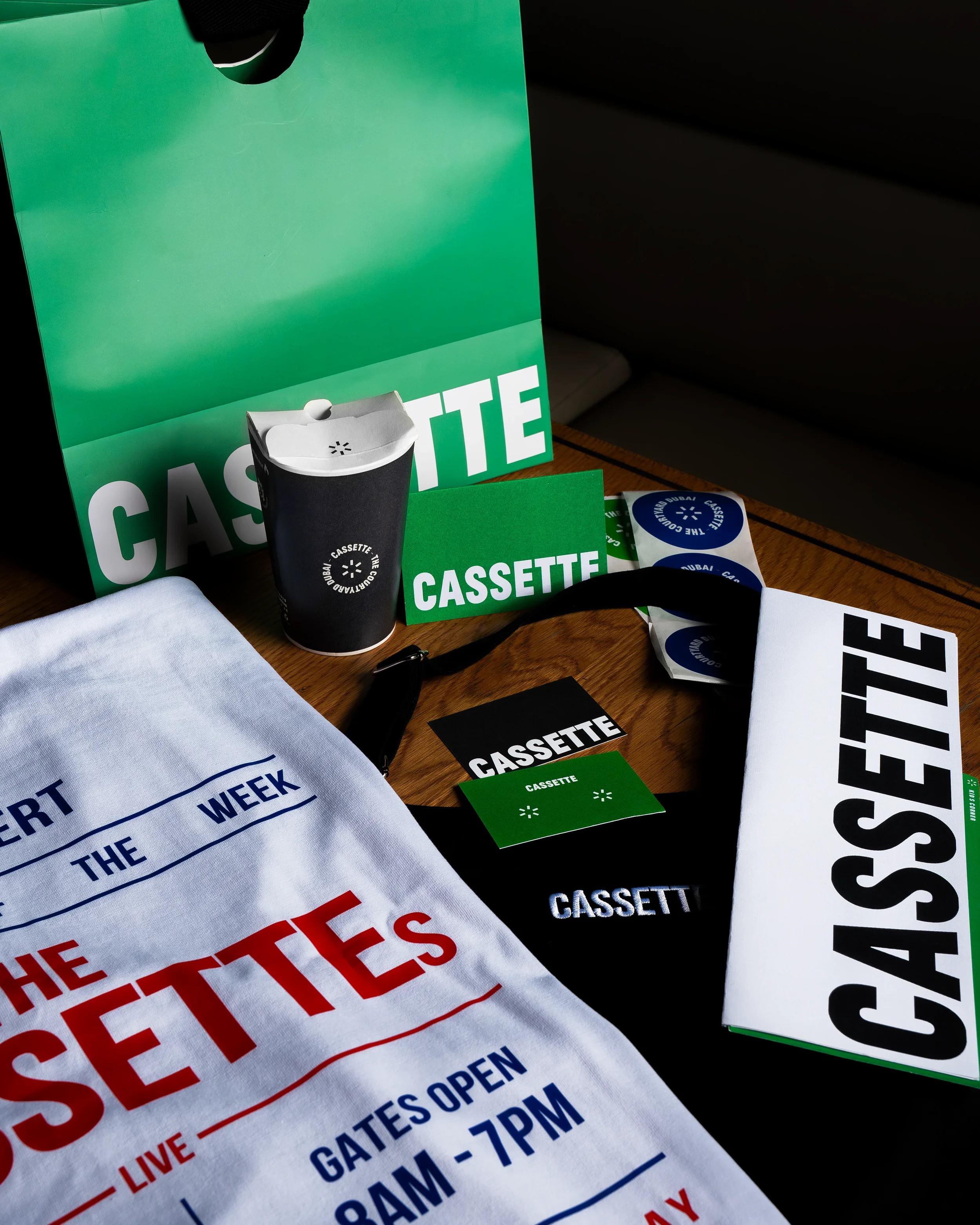 Close-up of various event-related printed materials on a wooden surface, including a black paper cup, green and white cassette-themed cards, a white cloth with red and blue text, and several circular stickers.