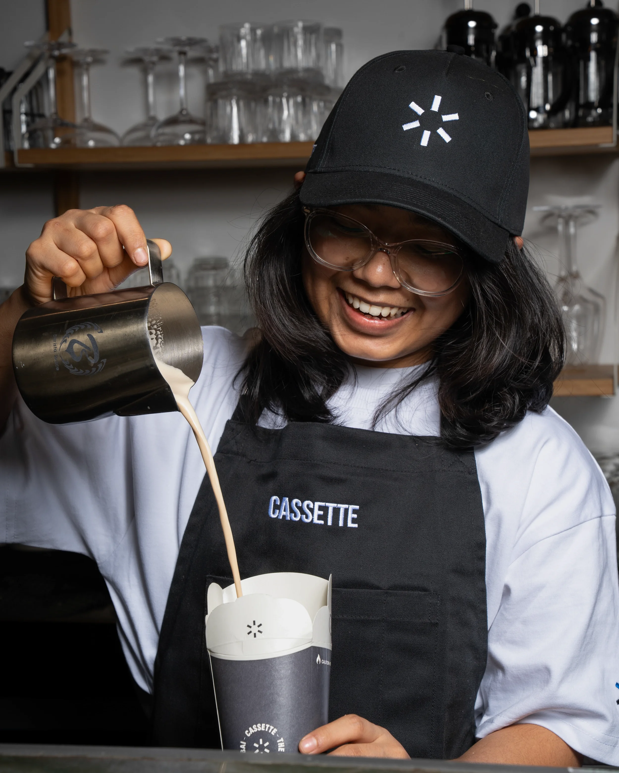 A woman wearing glasses and a black cap with a white loading symbol, smiling as she pours a drink from a metal shaker into a white cup with the word 'CASSETTE' on it. She is wearing a white shirt and a black apron.