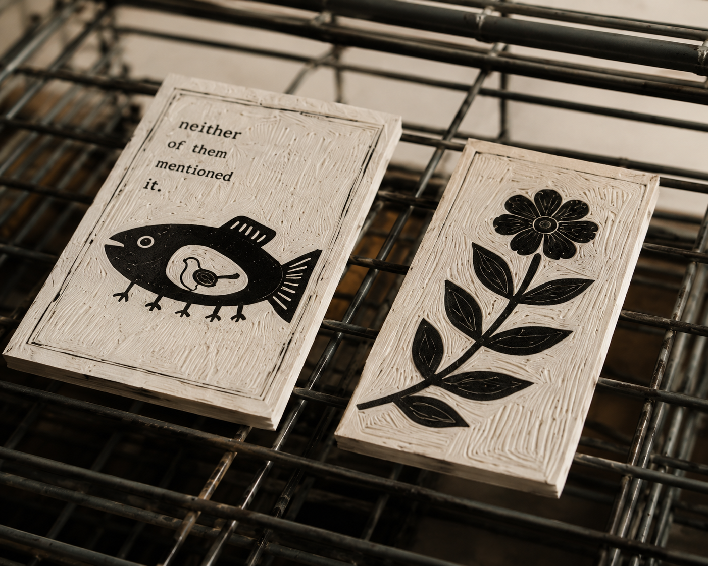 Two art prints with black illustrations and text on a wire rack. The left plaque has a stylized fish with a small bird inside and the text 'neither of them mentioned it.' The right plaque has a black silhouette of a flower and leaves.