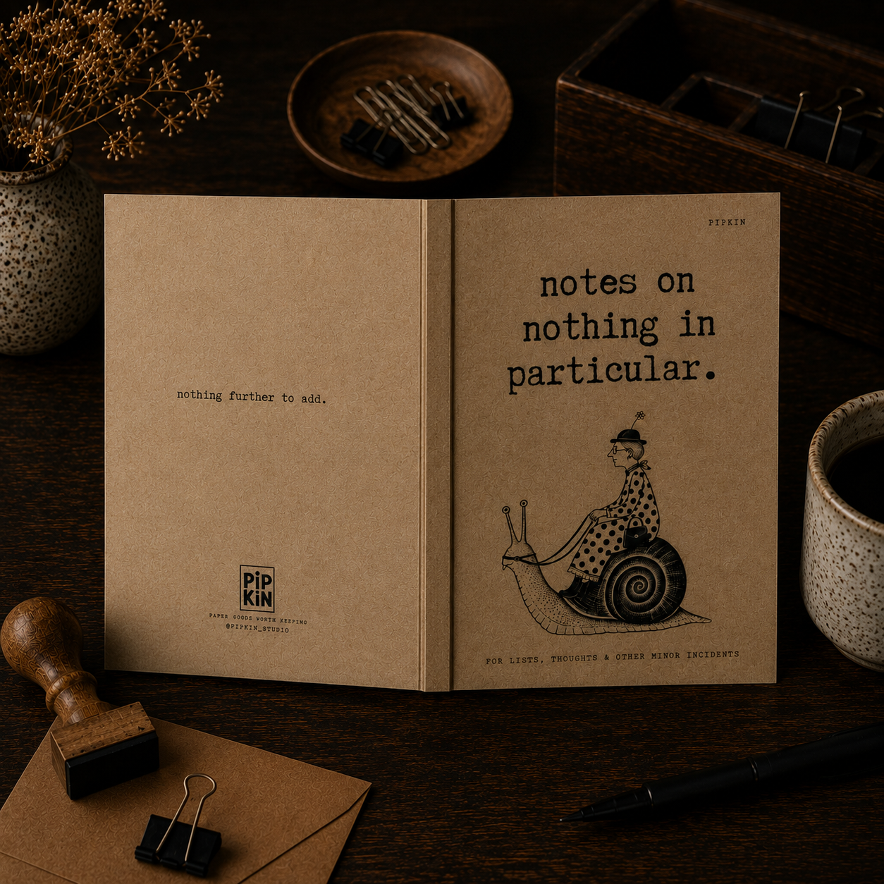 Open notebook with the phrase 'notes on nothing in particular' and a drawing of a woman riding a snail. The dark wooden desk has a cup of coffee, a small bowl with paper clips, another bowl with pins, a rubber stamp, a black pen, and decorative dried flowers in a speckled vase.