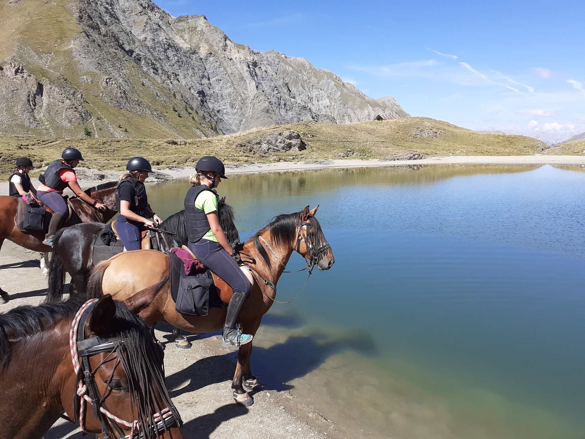 Groupe de personnes à cheval au bord d'un lac de montagne, avec des randonneurs portant casques et gilets de sécurité, entouré de montagnes rocheuses et d'un ciel bleu.