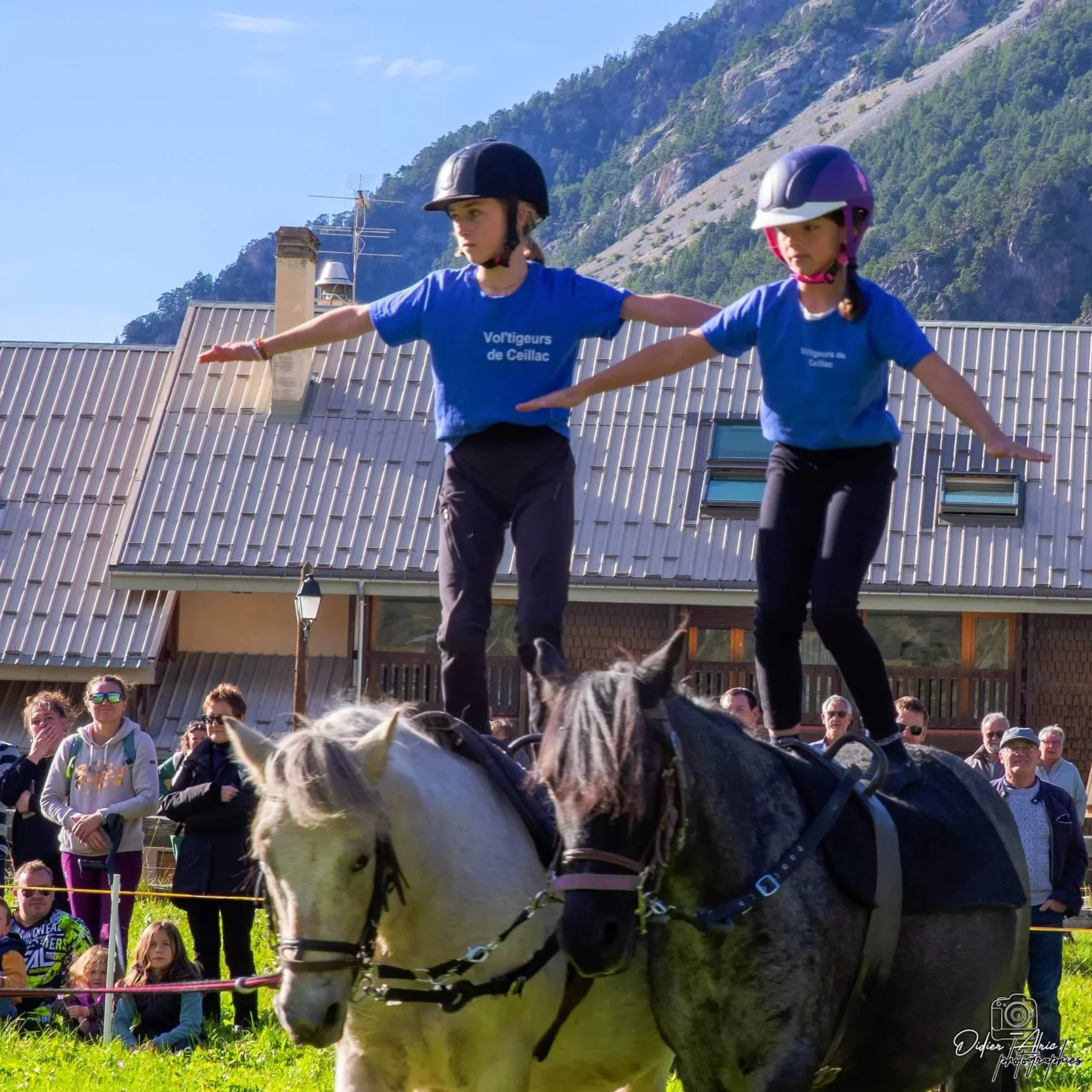 Deux jeunes femmes en casque de roller ou skateboard, portant des t shirts bleus, font du roller ou skateboard, entourées de spectateurs, avec une montagne en arrière-plan.
