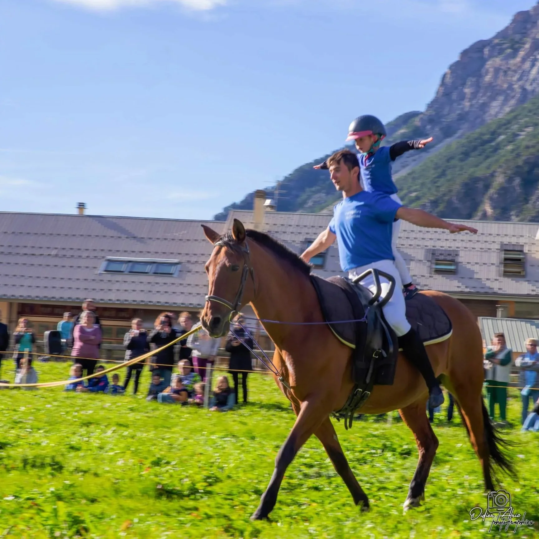 Un homme portant un casque rouge et un enfant sur ses épaules, avec des bras tendus, faisant du cheval à un événement hippique, avec un bâtiment et des montagnes en arrière-plan et un groupe de spectateurs au premier plan.