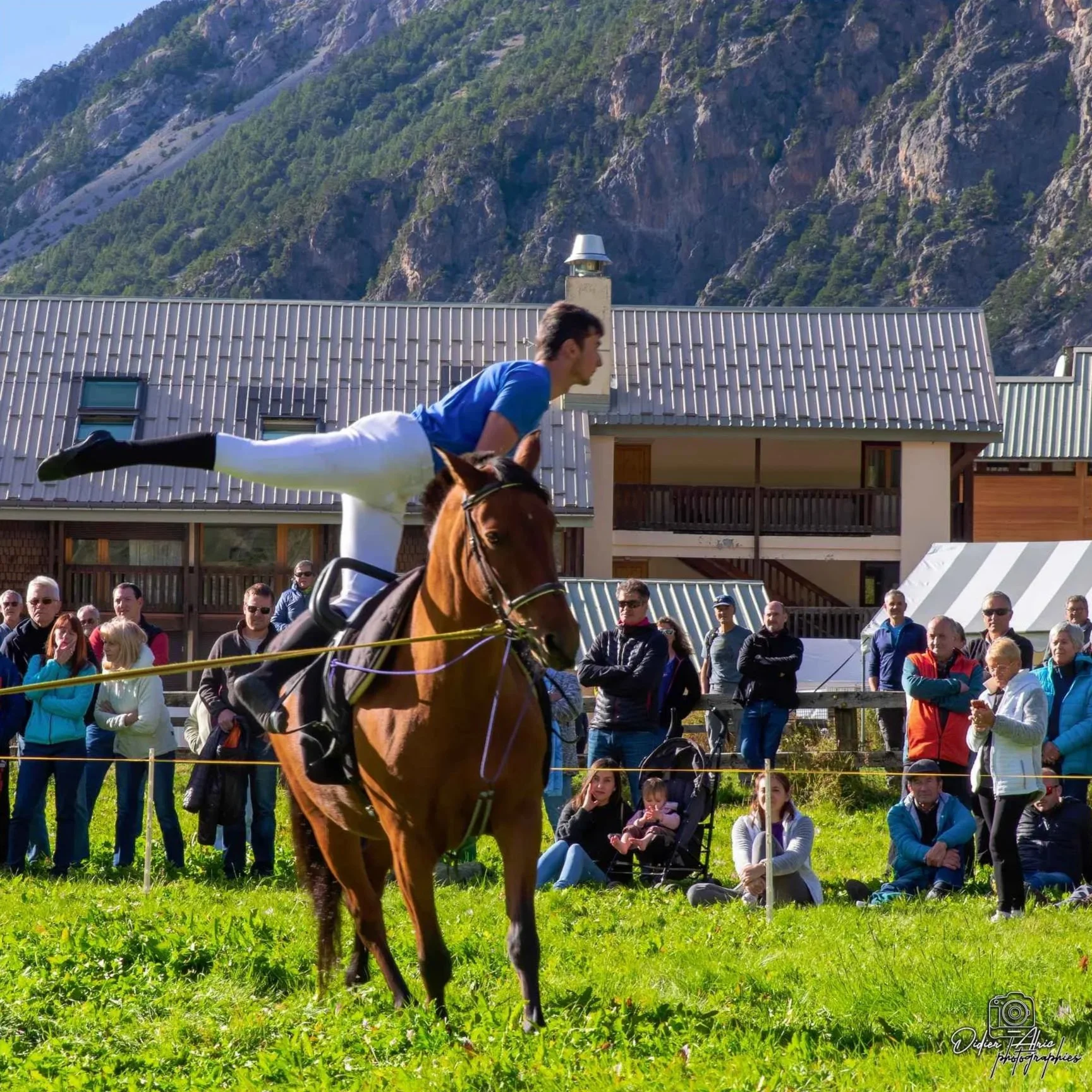 Une course de chevaux avec un jockey en bleu en avant-plan et un public regardant dans un jardin avec une montagne en arrière-plan.