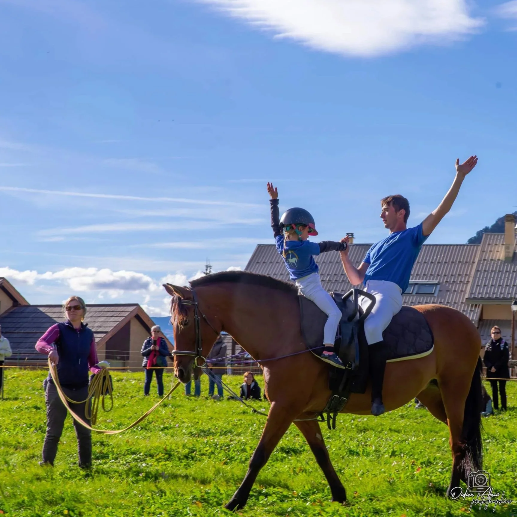 Personne à cheval en plein air, avec deux enfants montant également à cheval et une femme à côté, dans un environnement de campagne ensoleillée.