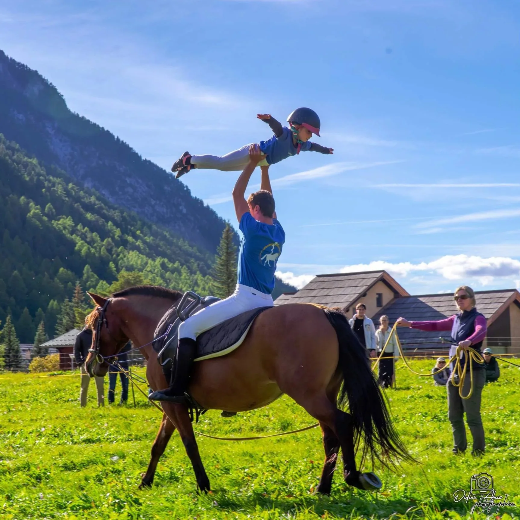 Un enfant en casque de roller est tenu en l'air par une personne à cheval, dans un champ vert avec une montagne en arrière-plan et un ciel bleu clair.