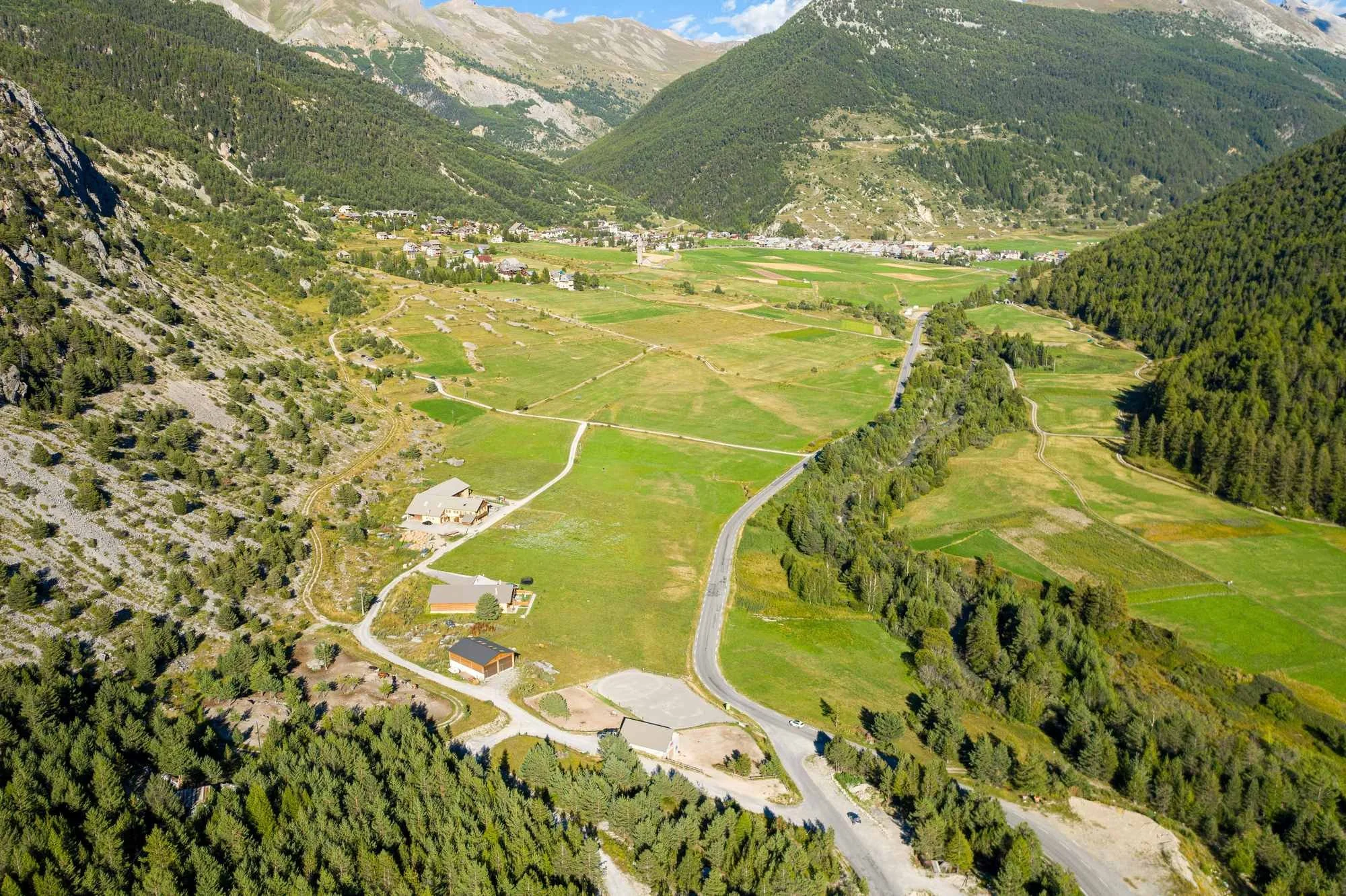 Vue aérienne d'une vallée verdoyante encerclée par des montagnes, avec des champs agricoles, des routes sinueuses et quelques bâtiments résidentiels.