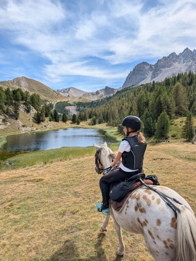 Une jeune fille en casque et gilet de protection à cheval dans un paysage de montagne avec un lac et des arbres.