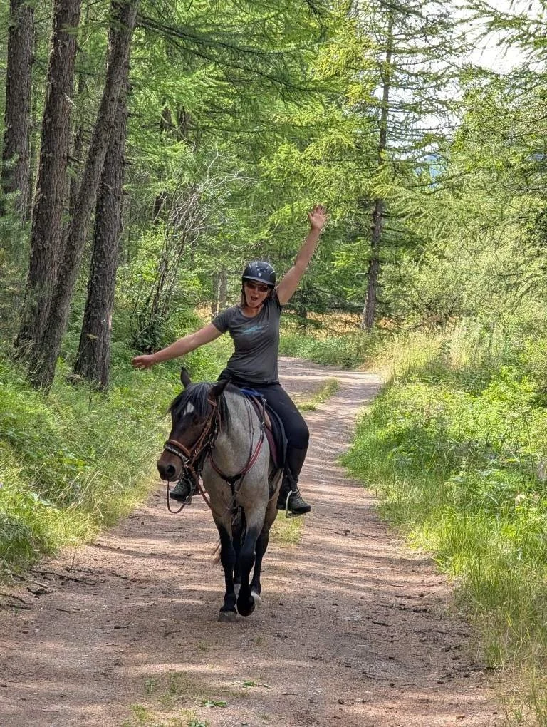 Une femme souriante portant un casque, assise sur un cheval gris, traverse un sentier forestier en plein été, avec des arbres verts et un ciel clair