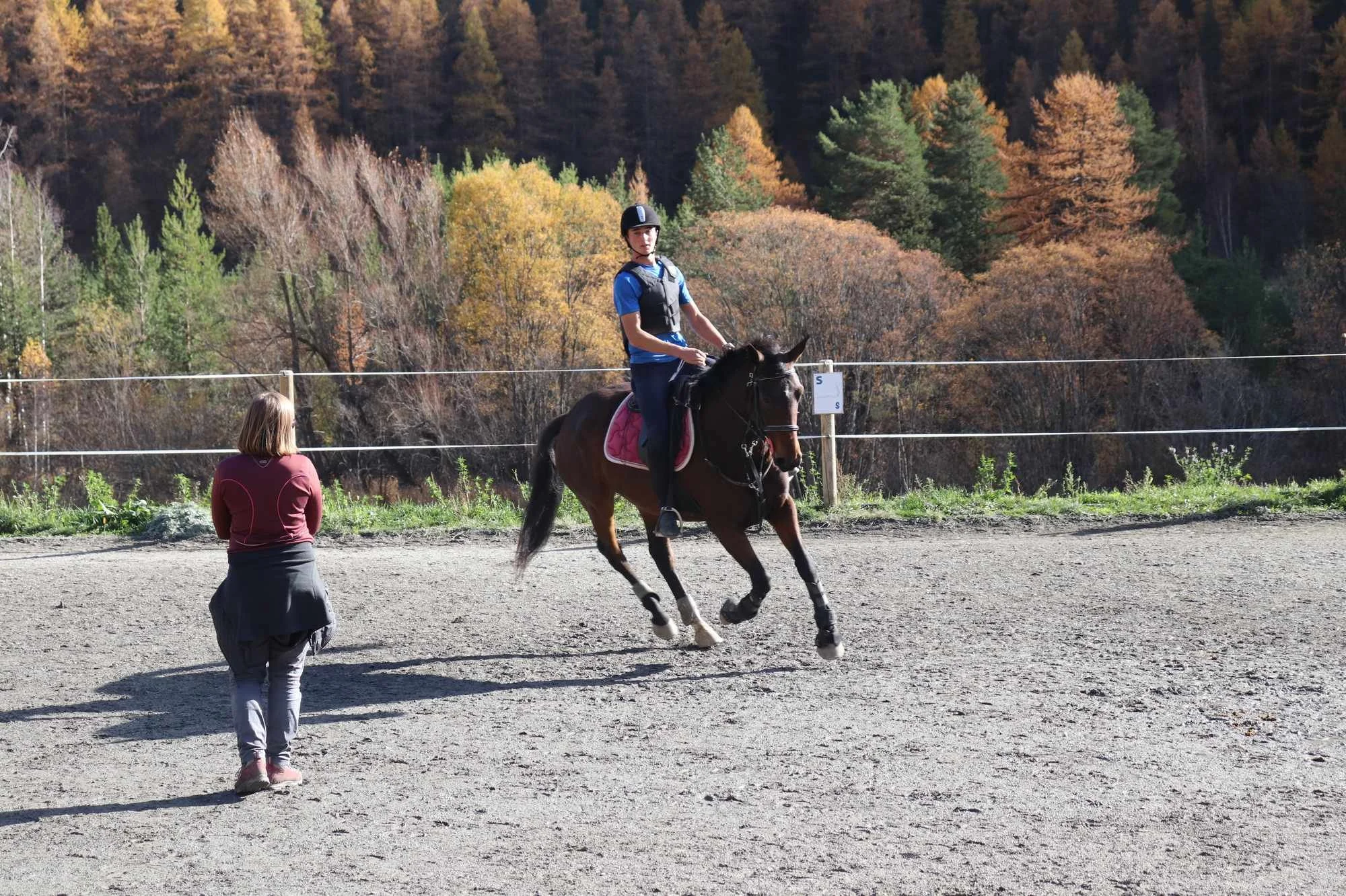Un jeune homme en casque et gilet de sécurité monte à cheval dans une zone extérieure, une femme le regarde et une autre personne en chemise rouge et pantalon clair se trouve en face. Le décor comprend des arbres automnaux dans les montagnes.