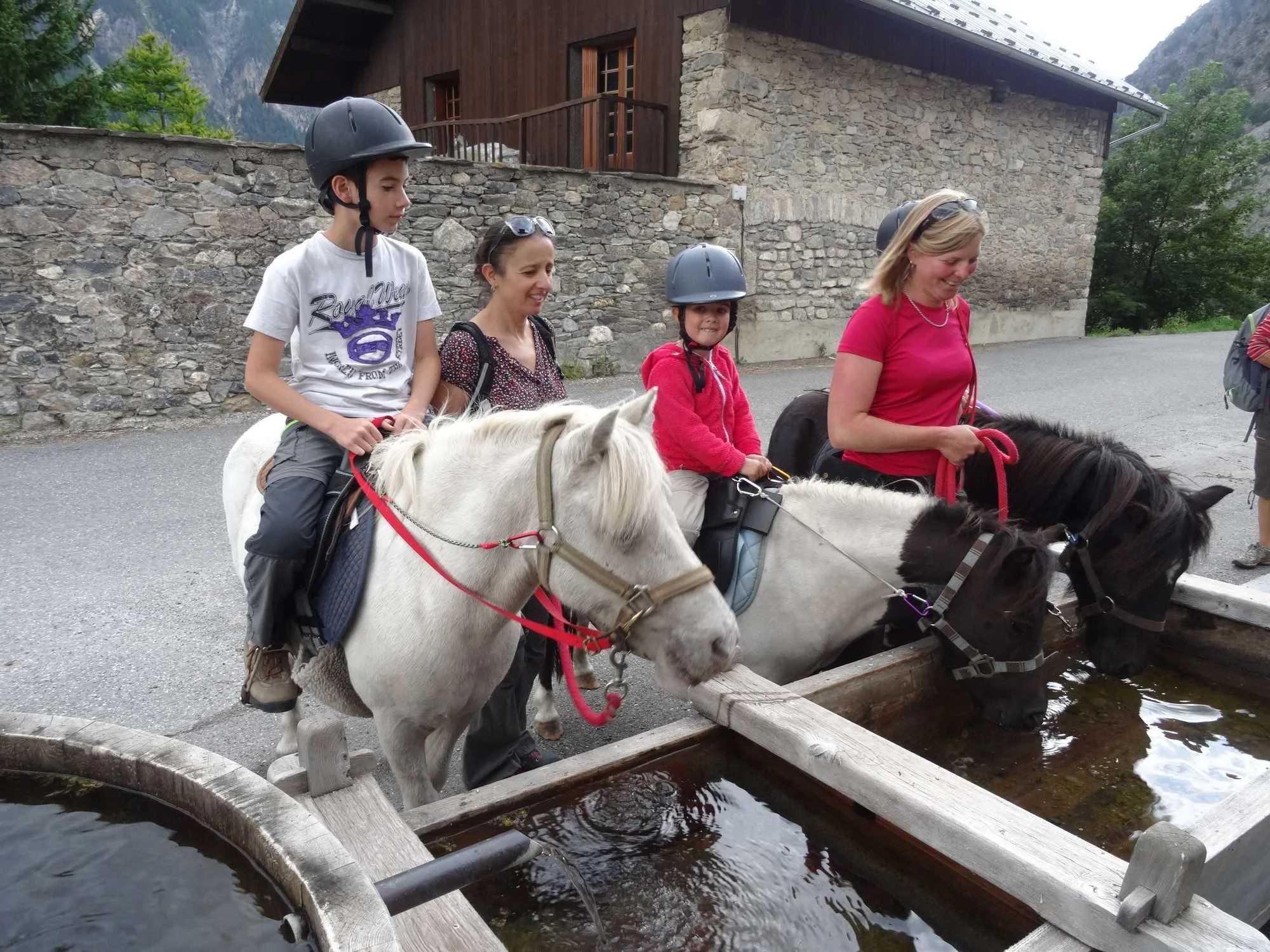 Groupe de personnes, y compris des enfants, chevauchant des poneys près d'un plan d'eau, avec une cabine en bois et des montagnes en arrière-plan.