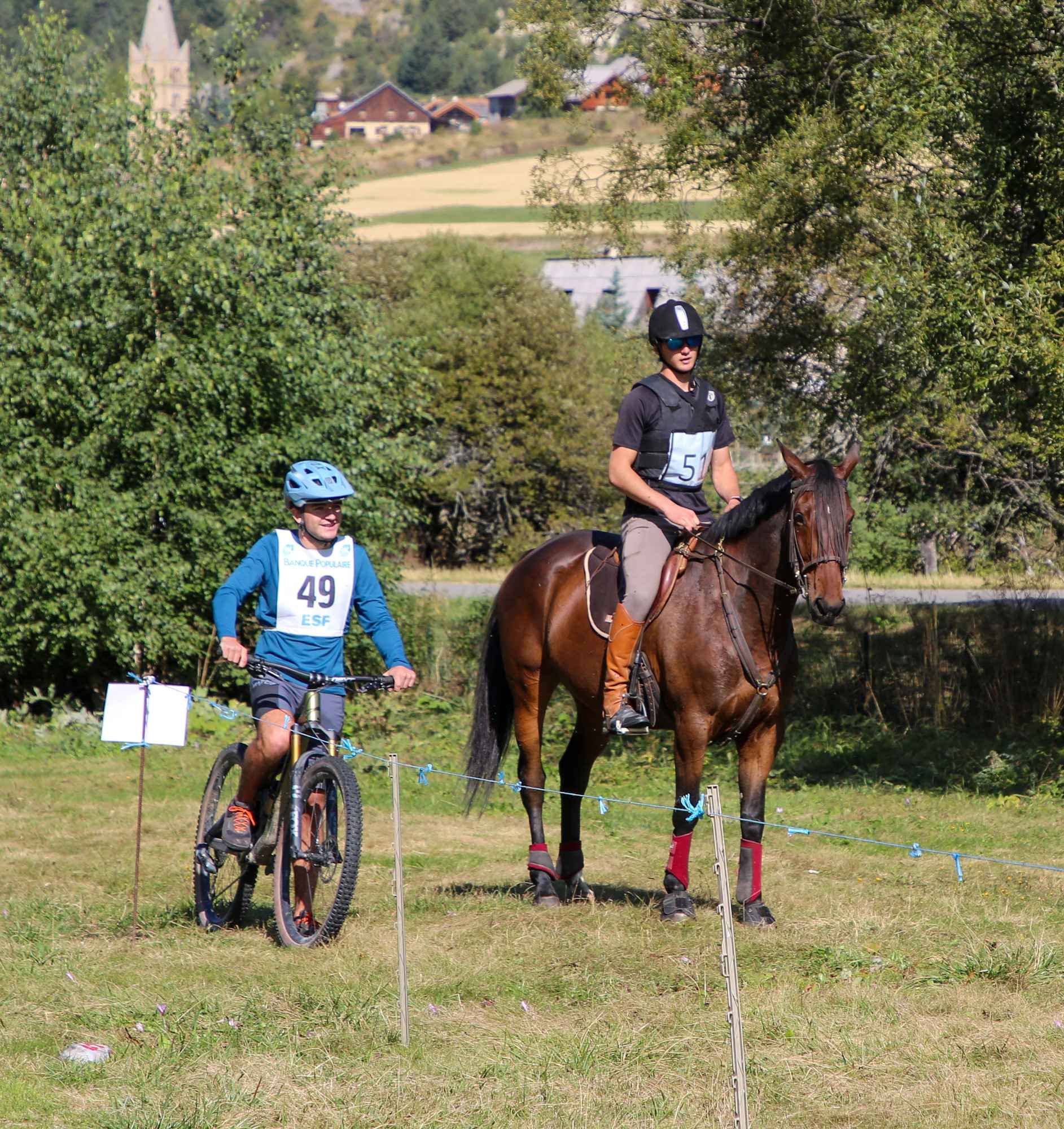 Deux personnes participent à une compétition, une à vélo et l'autre à cheval, dans un environnement naturel avec des arbres et des maisons en arrière-plan, portant des dossards avec des numéros.