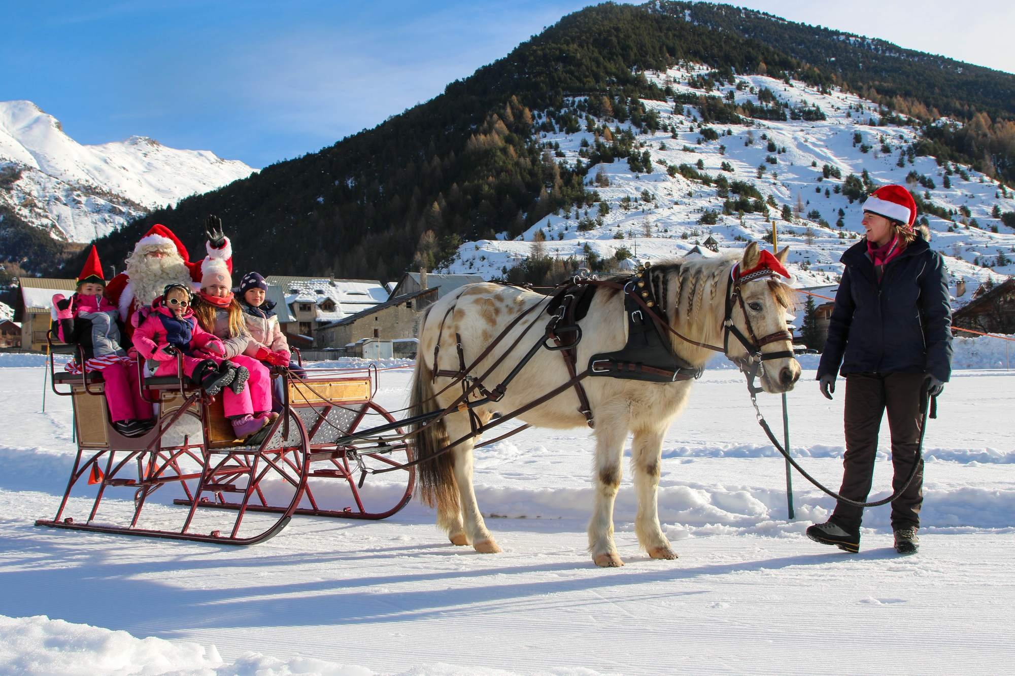 Enfant et adultes en traîneau avec Père Noël dans un paysage enneigé avec montagnes, personnes en vêtements d'hiver, père Noël saluant, femme avec un chapeau de Noël, et un cheval tirant le traîneau.