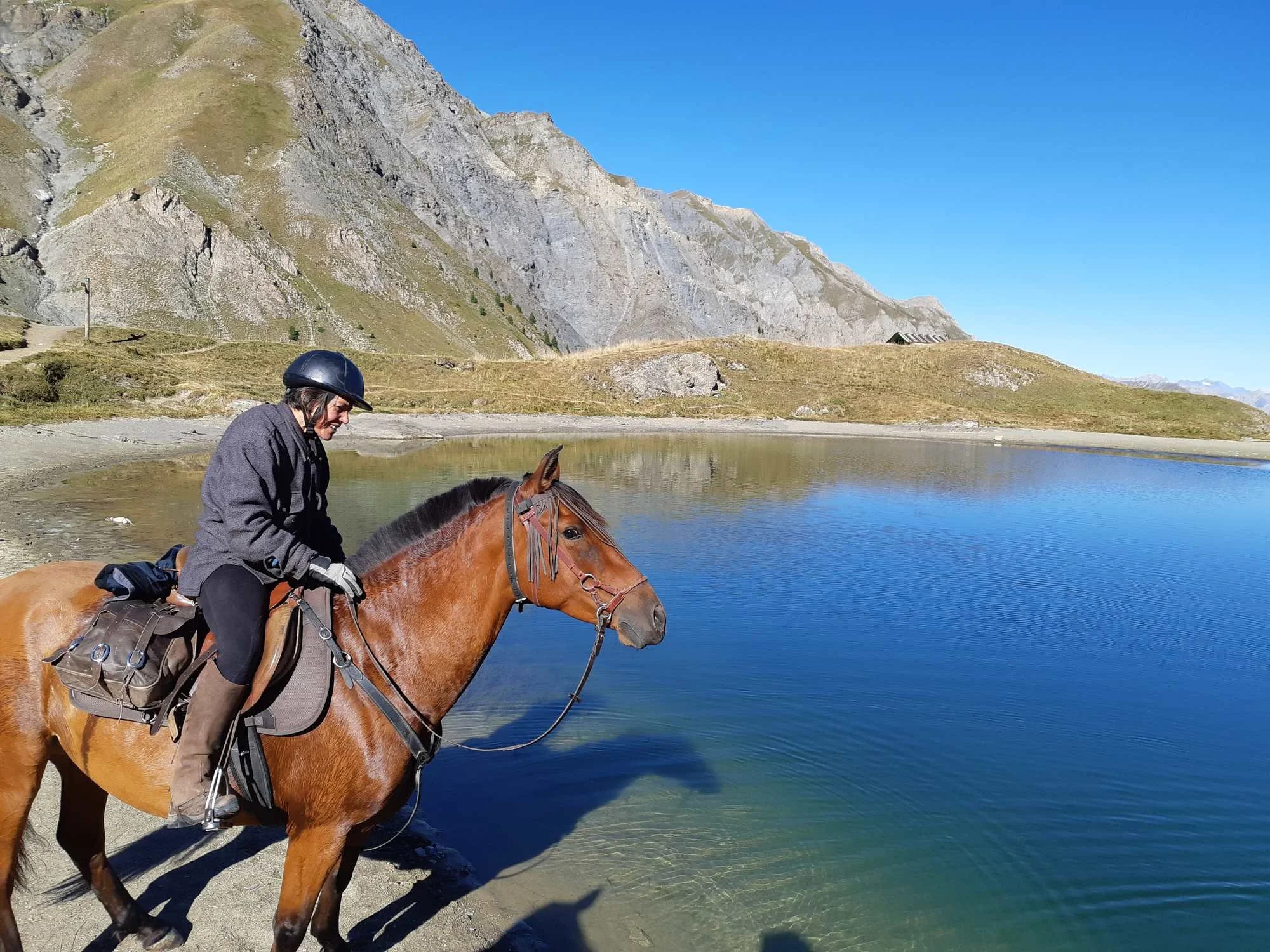Une personne à cheval près d'un lac dans un paysage montagneux, avec un ciel bleu clair.