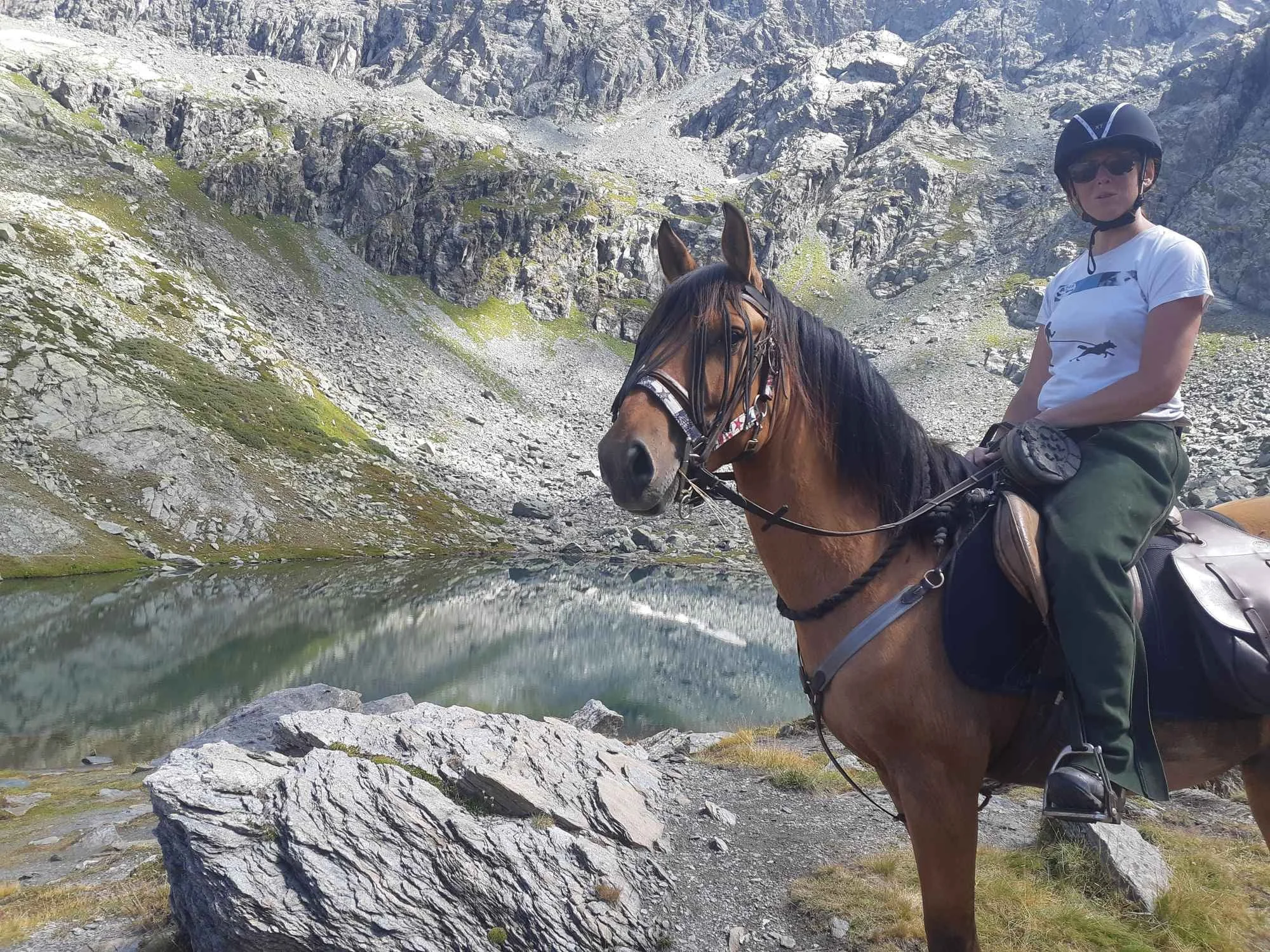 Une personne portant un casque et des lunettes de soleil, assise à cheval dans un paysage de montagne rocheux avec un lac en premier plan.