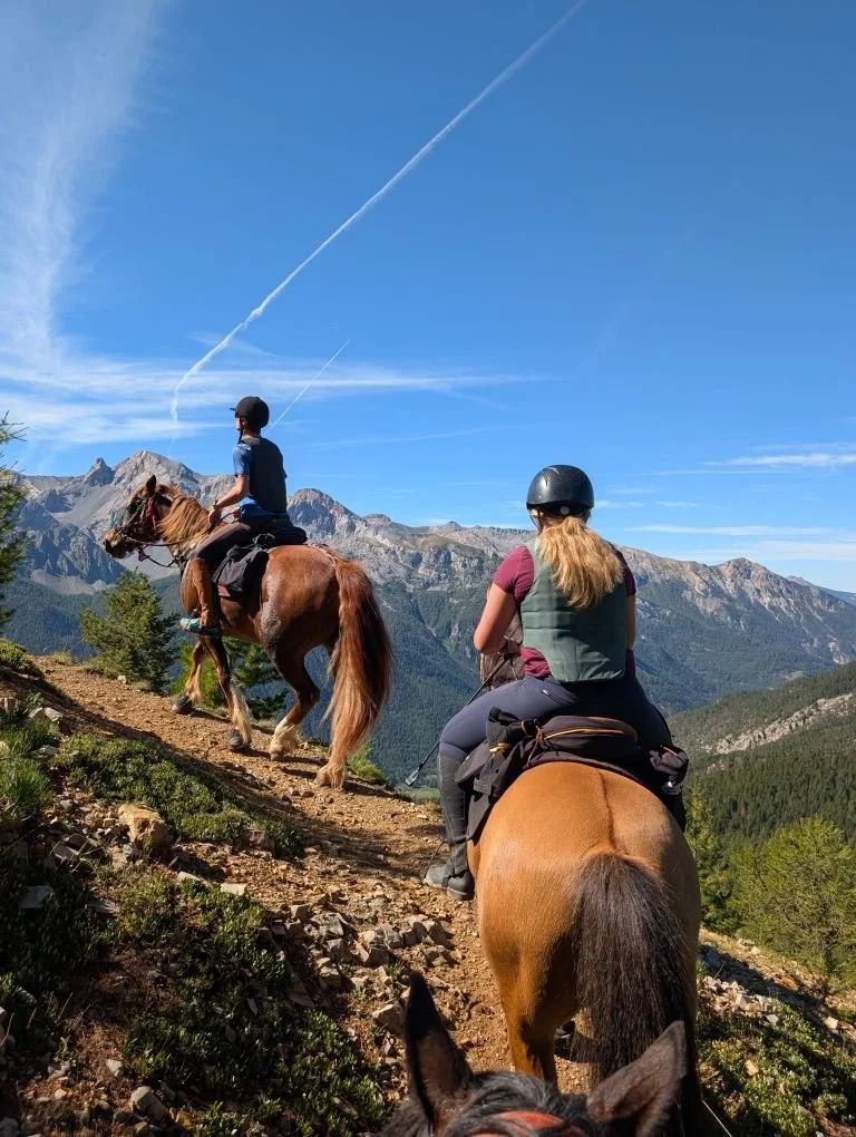 Deux personnes font de l'équitation en montagne, portant des casques et une veste de sécurité, avec des chevaux bruns sur un sentier rocheux, sous un ciel bleu avec des nuages et des traînées d'avion.