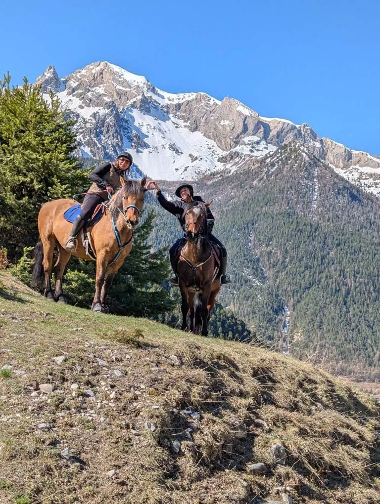 Deux personnes à cheval sur un terrain de montagne, avec une chaîne de montagnes enneigées en arrière-plan, un arbre vert à gauche.