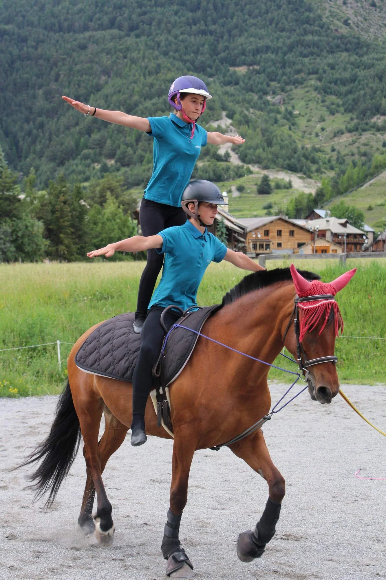 Deux jeunes cavaliers, un garçon et une fille, portent un casque et des habits bleus, et sont assis sur un cheval marron avec un tapis noir. La fille pratique la pose du fan ou T-pose sur le cheval, avec ses bras tendus horizontalement. Le décor en a