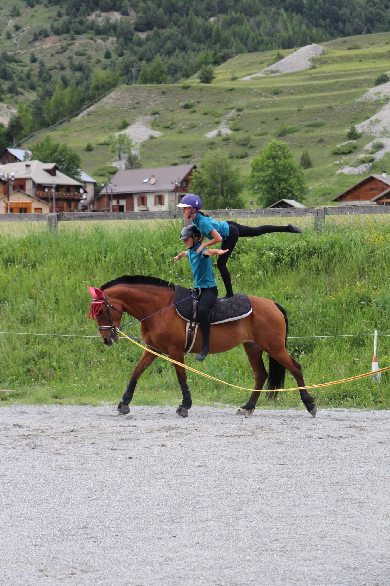 Deux personnes faisant de l'équitation, une personne moins âgée derrière une plus jeune, portant des casques, dans un environnement rural avec des maisons en bois et des collines verdoyantes.