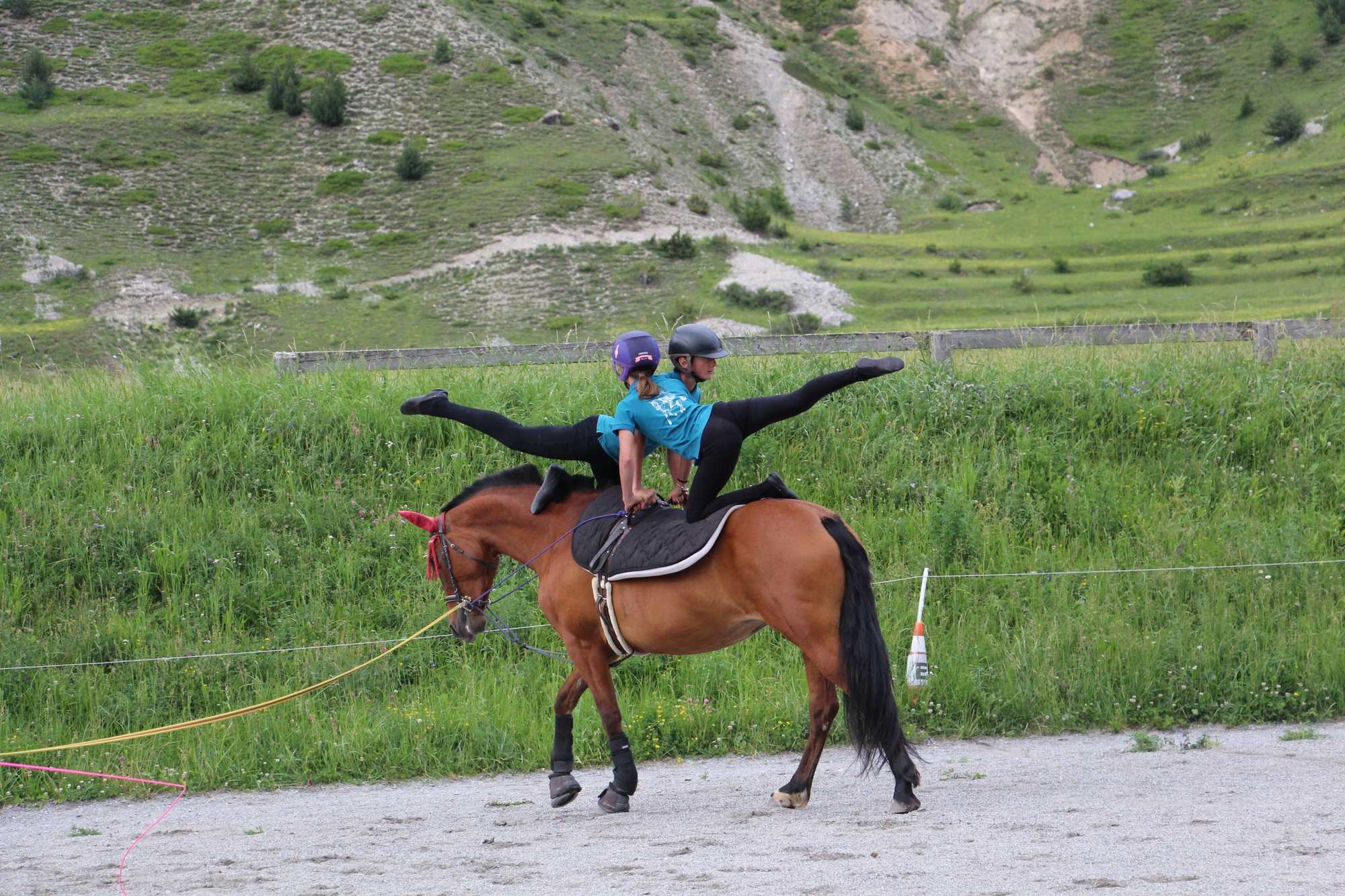 Deux personnes faisant de l'équitation dans un champs vert avec une montagne en arrière-plan. L'une d'elles est allongée sur le dos du cheval, en équilibre sur une jambe, portant un casque et une veste bleue, tandis que la personne en dessous porte é