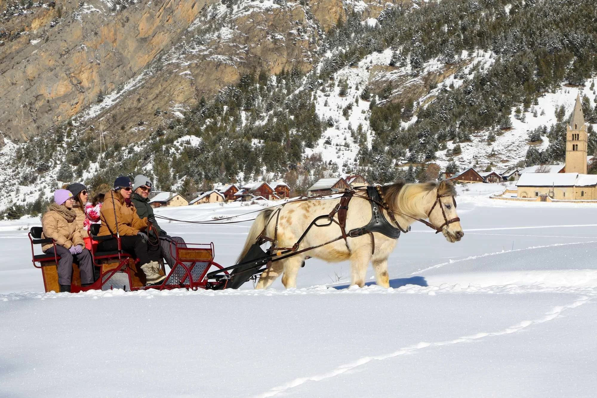 Groupe de personnes faisant une promenade en traîneau à cheval dans un paysage enneigé, avec des maisons en bois et une église au fond, dans un environnement montagnard.