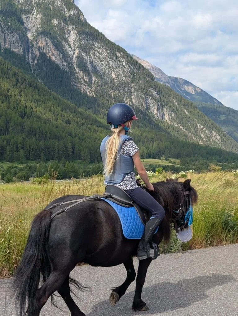 Une fille chevauche un poney noir avec un casque de protection dans un paysage montagneux avec des collines verdoyantes et un ciel partiellement nuageux.