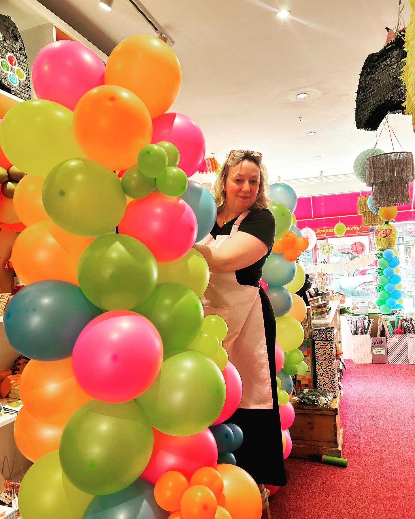 A woman standing behind a colorful balloon display inside a store. The balloons are various bright colors, including pink, orange, green, and blue, arranged in a large cluster. The store has shelves with various products and a pink wall with decorati
