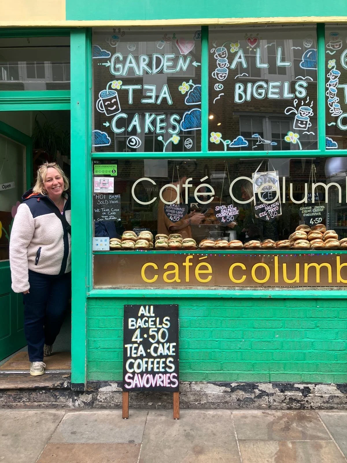 The storefront of Café Columbo with green framing, displaying various bagels and baked goods in the window. The window has colorful signs advertising garden tea, cakes, bigels, and other items. A woman stands to the left, smiling, next to a blackboard sign that lists bagels, tea, cake, coffees, and savories for $4.50 each.