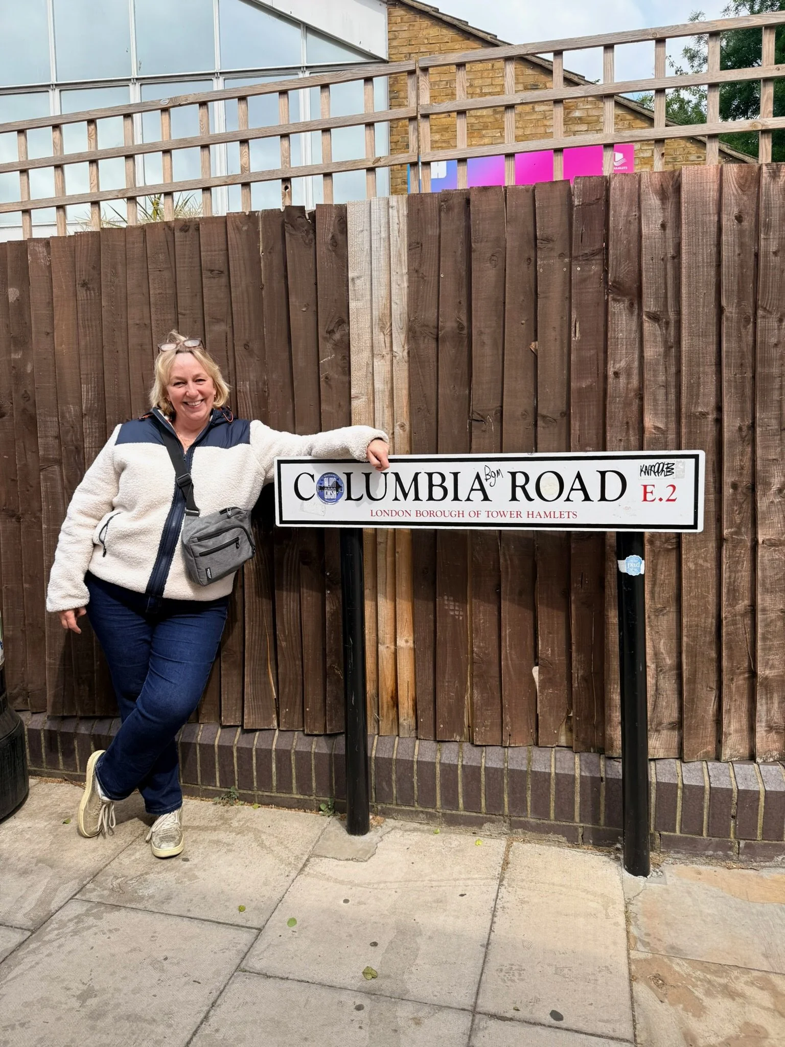 A woman smiling and posing next to a street sign that reads 'Columbia Road E.2,' with a wooden fence and a building with a glass window in the background.