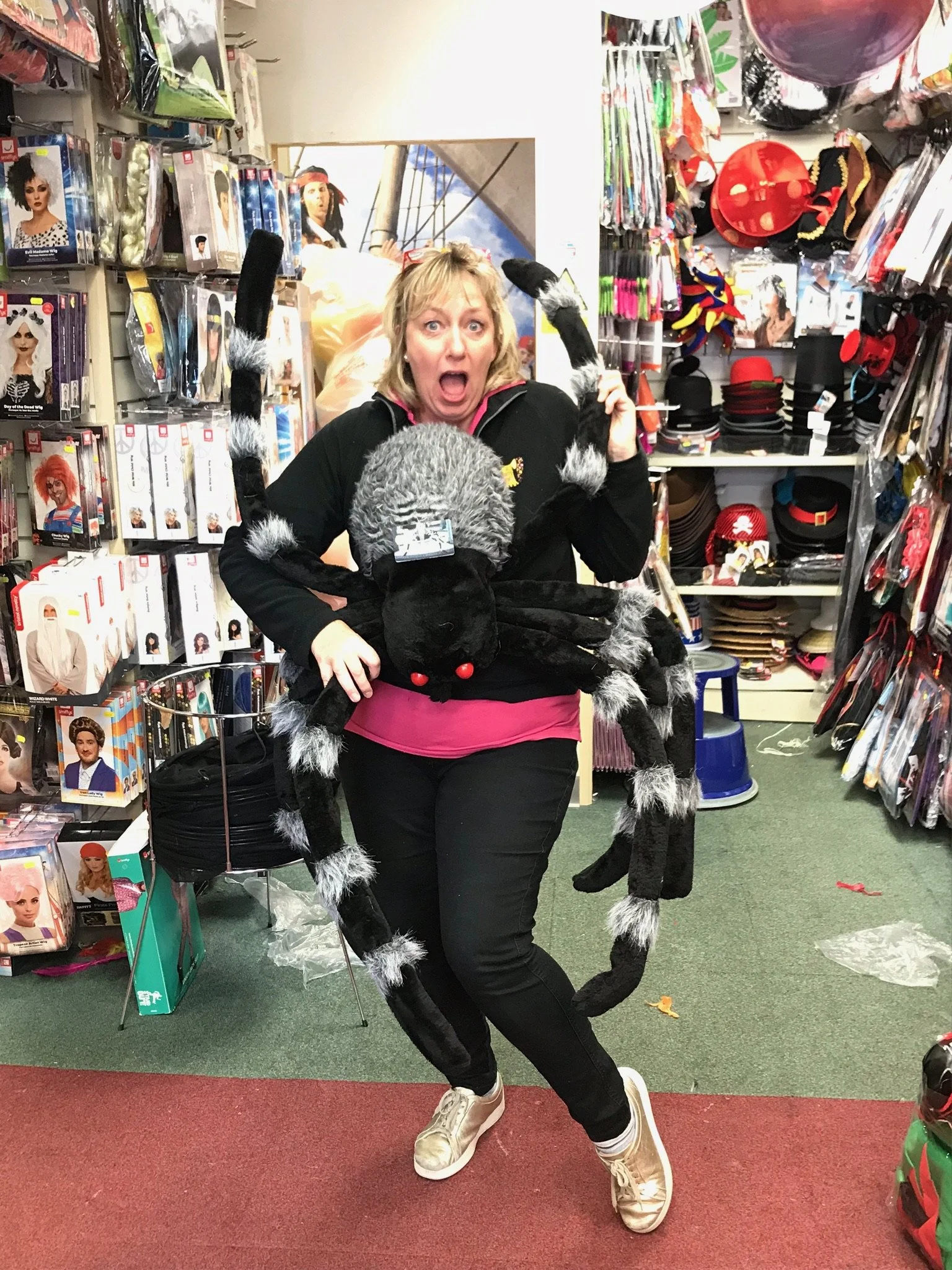 A woman dressed in a cat costume with black and white furry accents, hanging with a stuffed black cat toy, in a costume store aisle filled with wigs and hats.