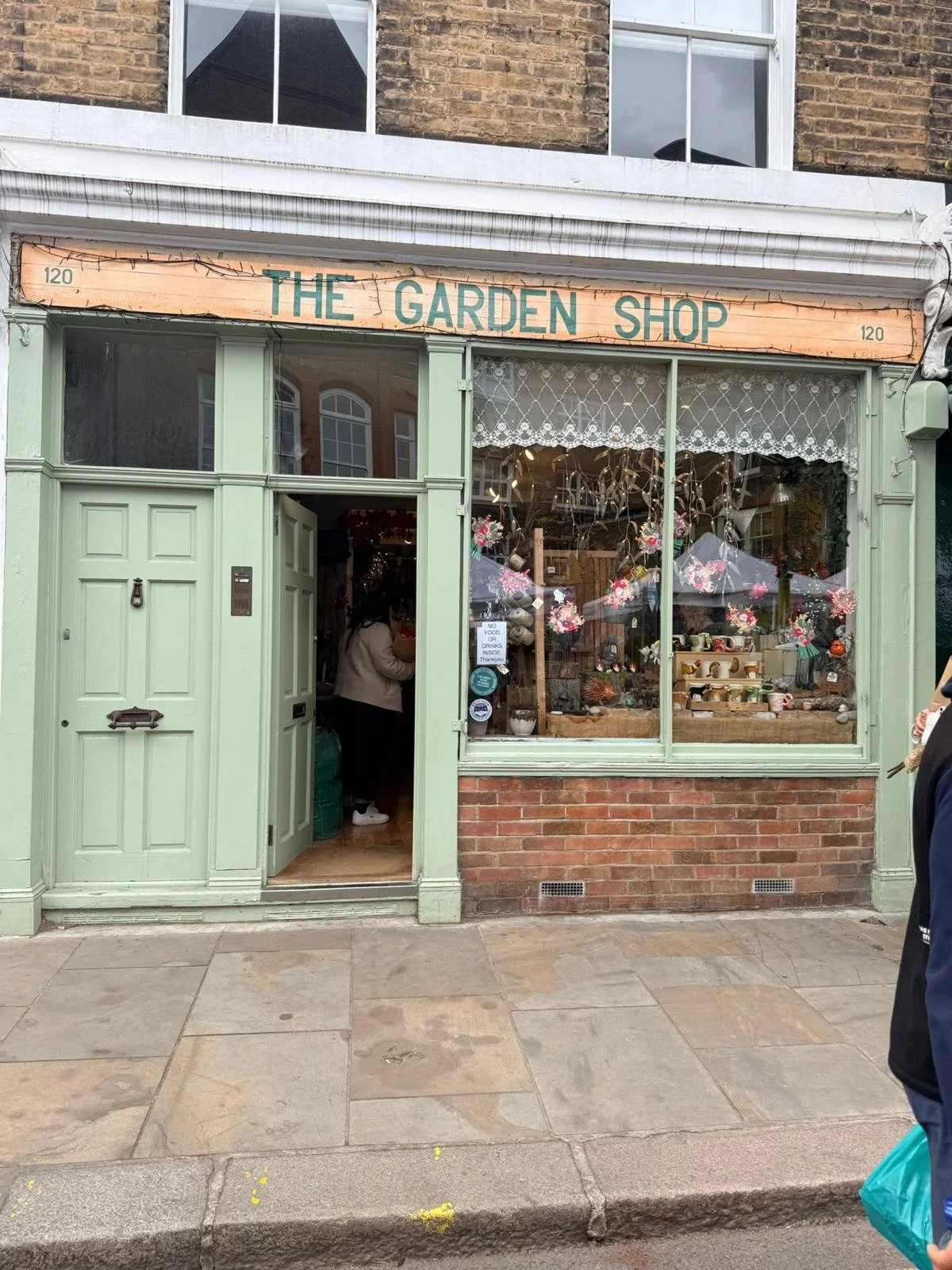 Front view of a shop called 'The Garden Shop' with a pastel green facade, large front window displaying floral decorations, and an open door showing customers inside.