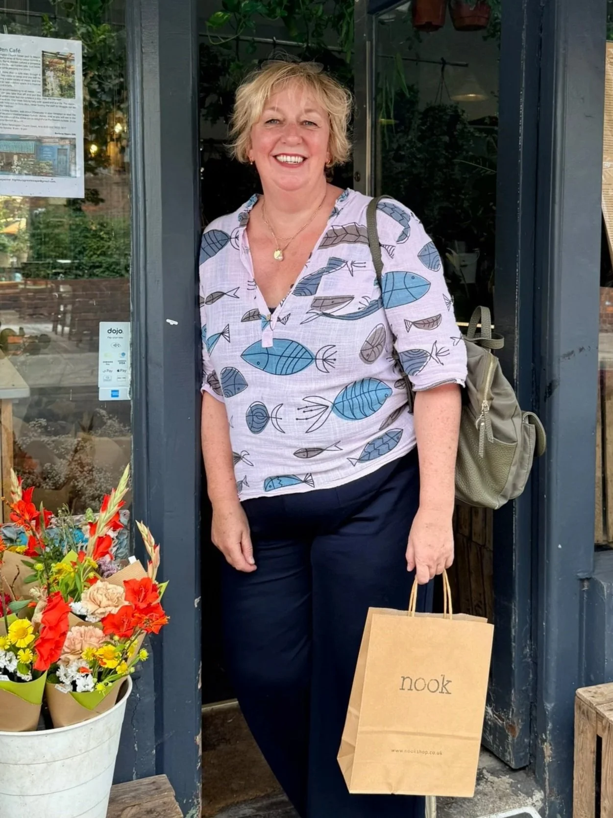 A woman standing at the entrance of a shop, smiling, wearing a fish-patterned shirt and carrying a beige backpack and a paper shopping bag labeled 'nook.' There is a colorful flower bouquet in a white pot at her side, and a menu or sign on the shop window behind her.